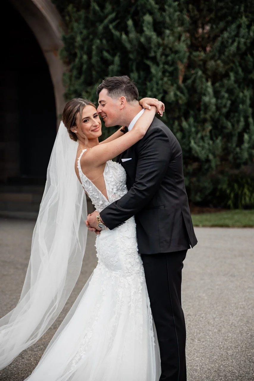 A bride and groom embracing outdoors on their wedding day, with the bride in a white wedding gown and veil and the groom in a black tuxedo. They are smiling and in a close, affectionate pose, with greenery in the background.