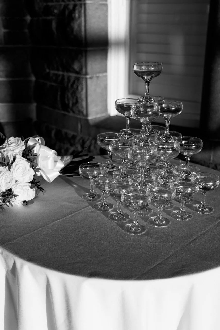 A champagne tower of glasses arranged in a pyramid on a table with a bouquet of white roses nearby, set against a brick wall and a wall with siding.