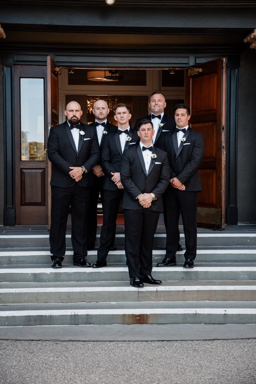 Six men dressed in black tuxedos with bow ties standing on steps outside a building.