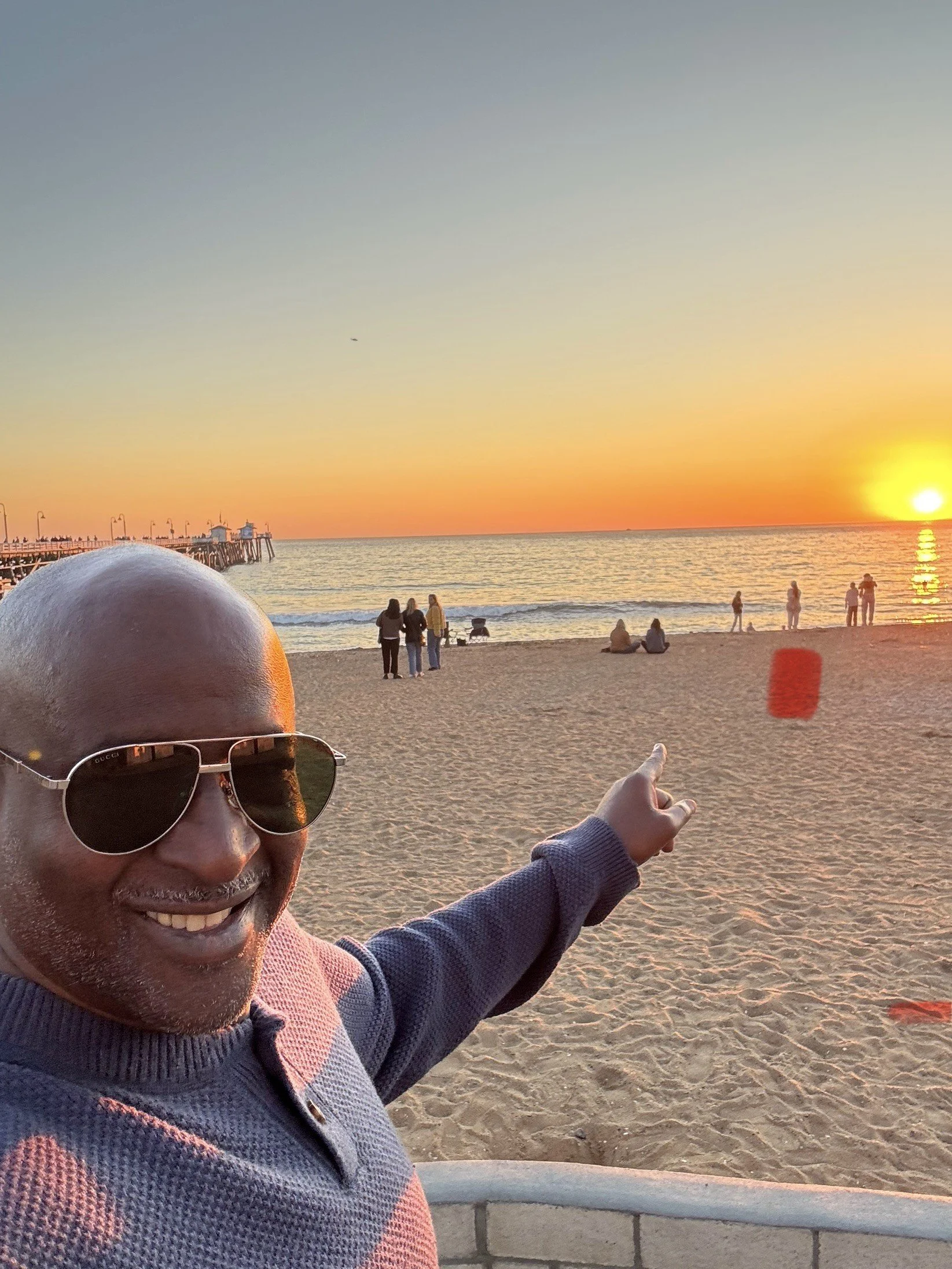 A smiling man wearing sunglasses and a sweater taking a selfie at the beach during sunset, pointing at the horizon with people in the background.
