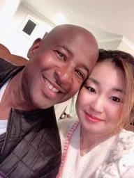 A man and a young woman taking a selfie together indoors. The man is smiling and the woman has a slight smile, both appearing happy.