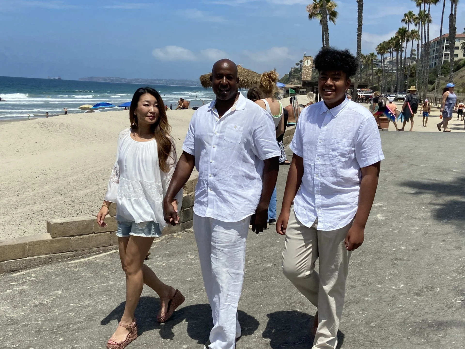 Three people walking on a beach with sand and ocean in the background. The woman on the left has long hair and is wearing a white top, denim shorts, and sandals. The man in the middle is bald, smiling, and wearing a white short-sleeved shirt and whit