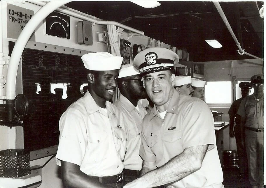 Black and white photo of a group of military personnel, including a man in a U.S. Navy uniform, in a control room with electronic equipment and a board with charts and numbers.