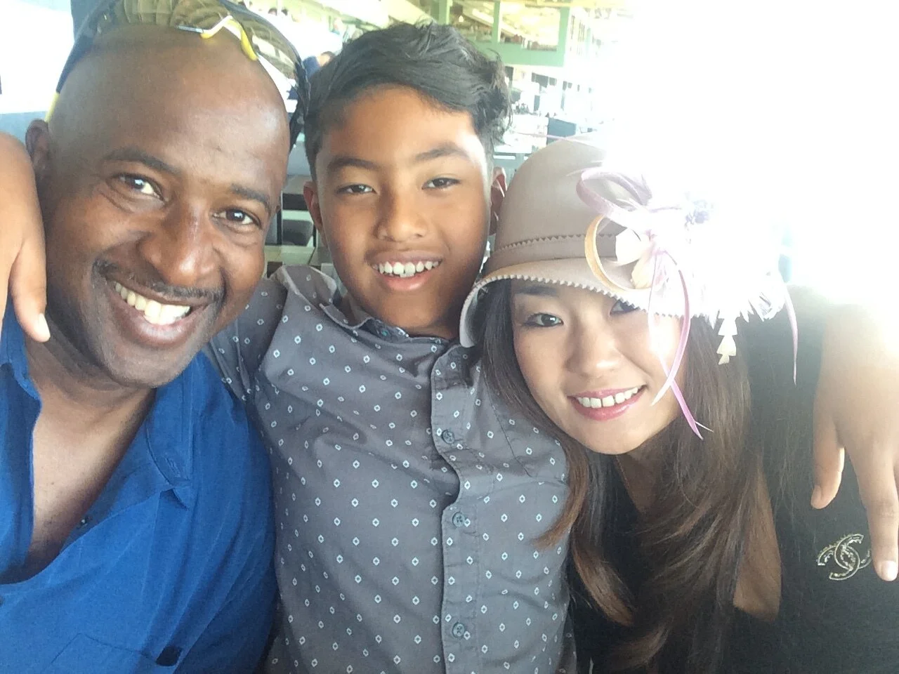 Three people smiling and posing closely together, with a bright background, one man and two children, one girl wearing a hat with feathers and ribbons.