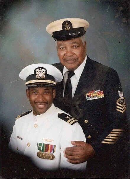 Two military personnel, one young woman and one older man, dressed in uniform with medals, posing together for a formal photo. The woman is wearing a white uniform with a peaked cap, and the man is in a dark uniform with a white cap. They are smiling