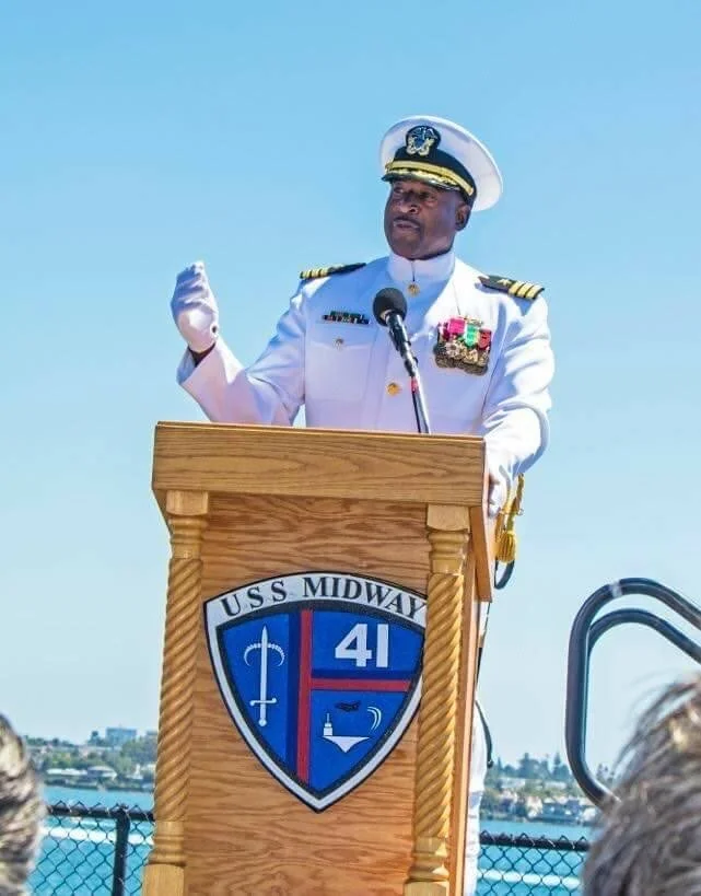 A man in a white naval uniform giving a speech at a podium outdoors, with a blue sky background. The podium has a shield with the text "U.S.S. MIDWAY 41" and nautical symbols.