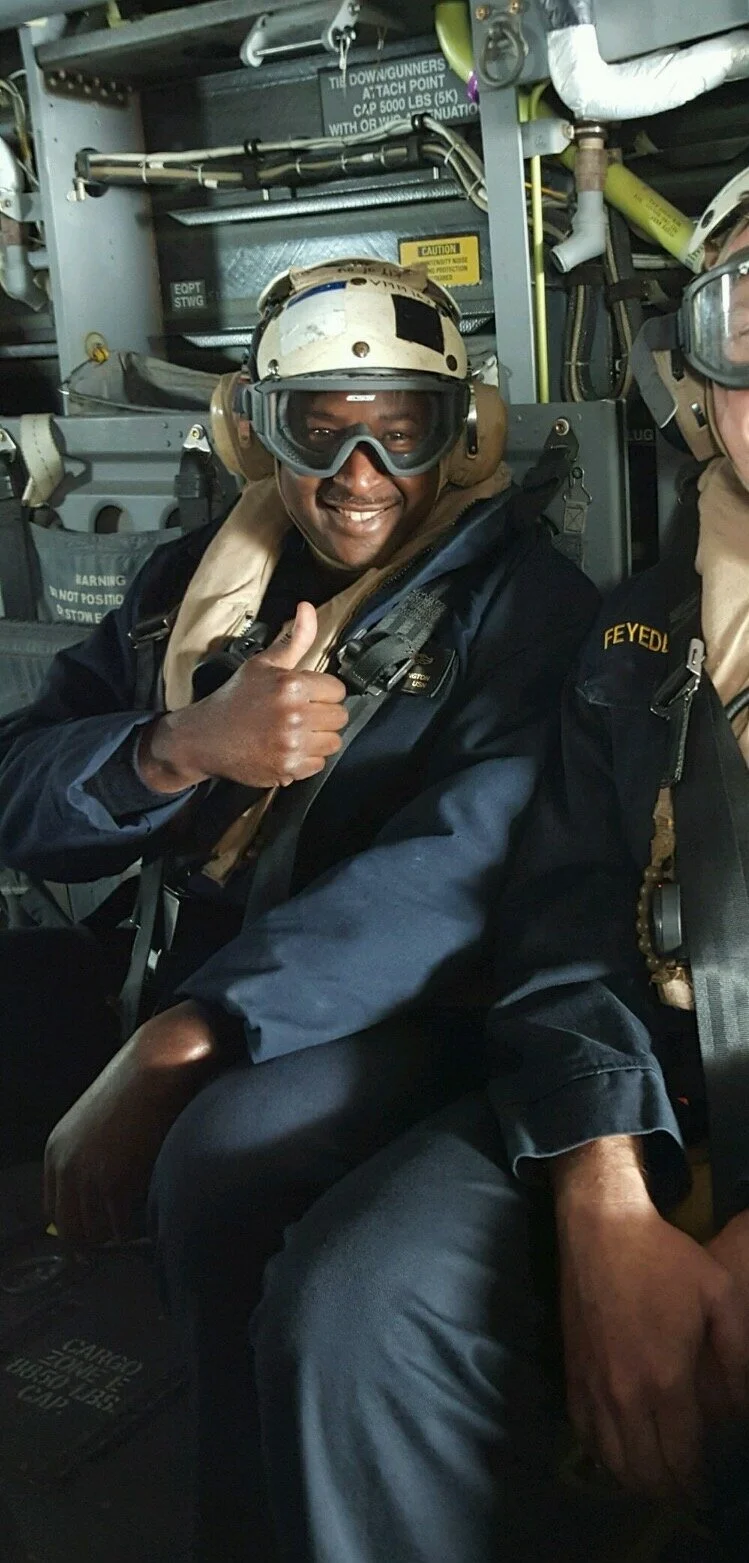 A smiling man in military or pilot gear, wearing a helmet and goggles, giving a thumbs up inside a helicopter or aircraft.