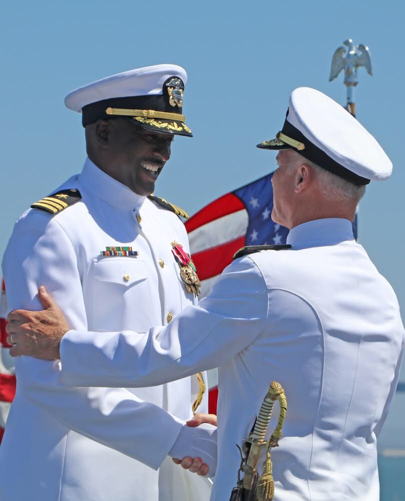Two uniformed military officers, exchanging a handshake and smiling against a backdrop of American flags and a clear blue sky.