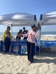 People at a beach booth with a sandy shoreline and ocean in the background, under white canopy tents.