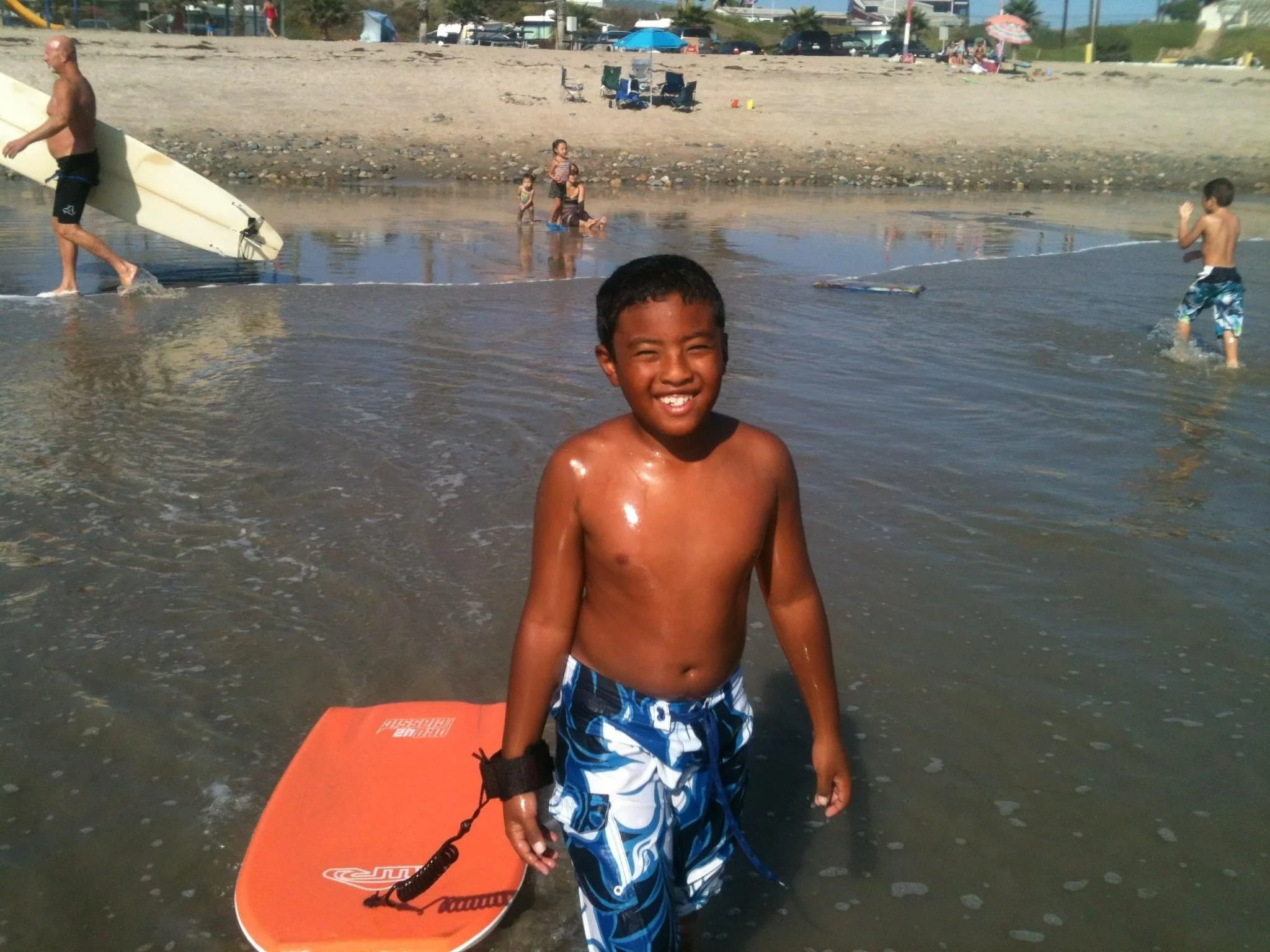 A young boy in blue swim trunks smiling while standing in shallow ocean water, holding a bright orange boogie board near a beach with other children and adults swimming, sunbathing, and carrying a surfboard in the background.