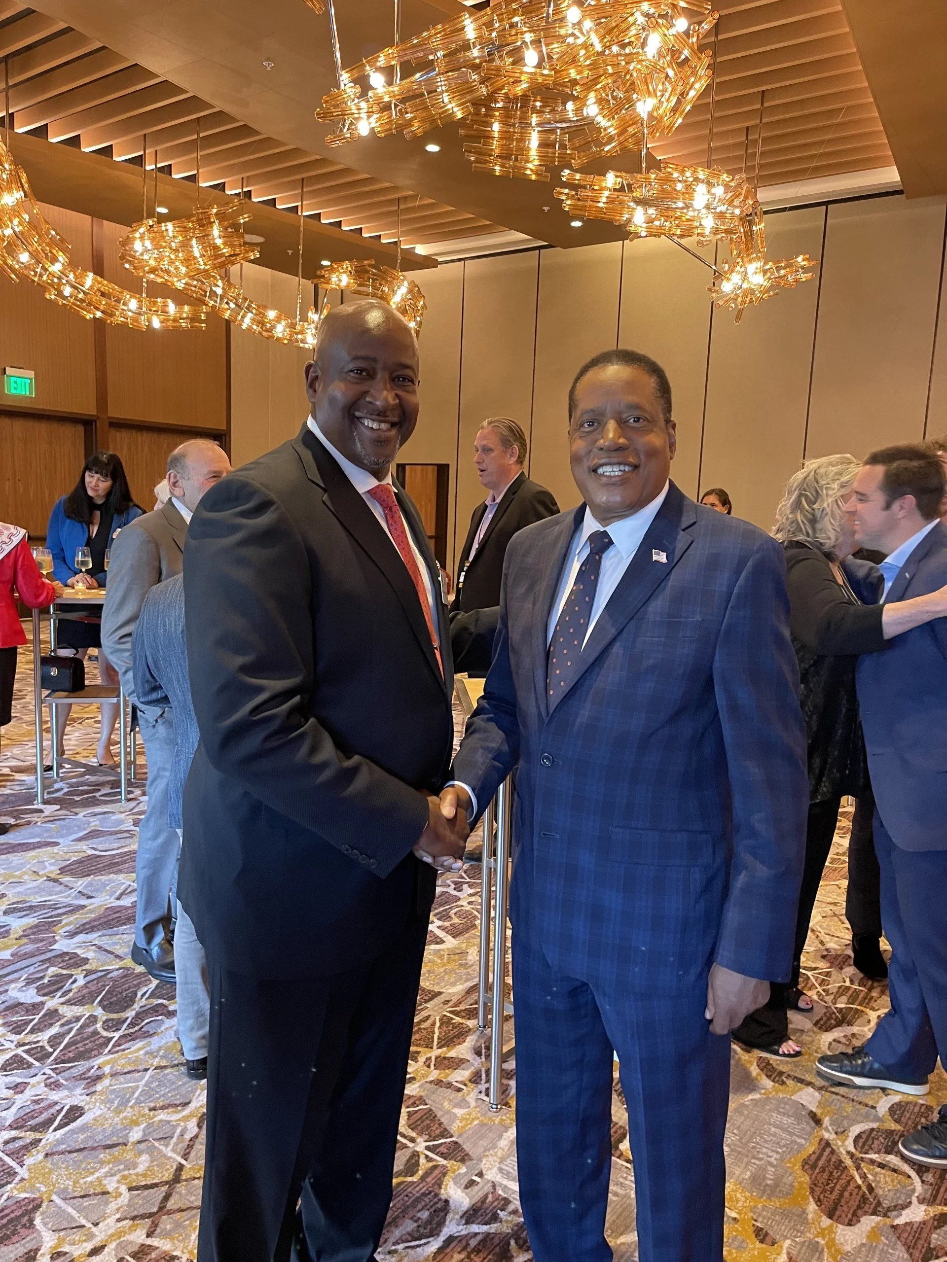 Two men in suits shaking hands at a formal event, with group of people in the background.
