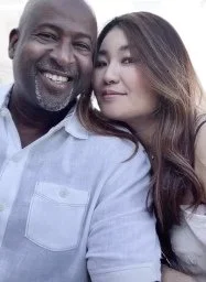 Close-up selfie of a smiling Black man and a young woman with long dark hair, both looking at the camera.