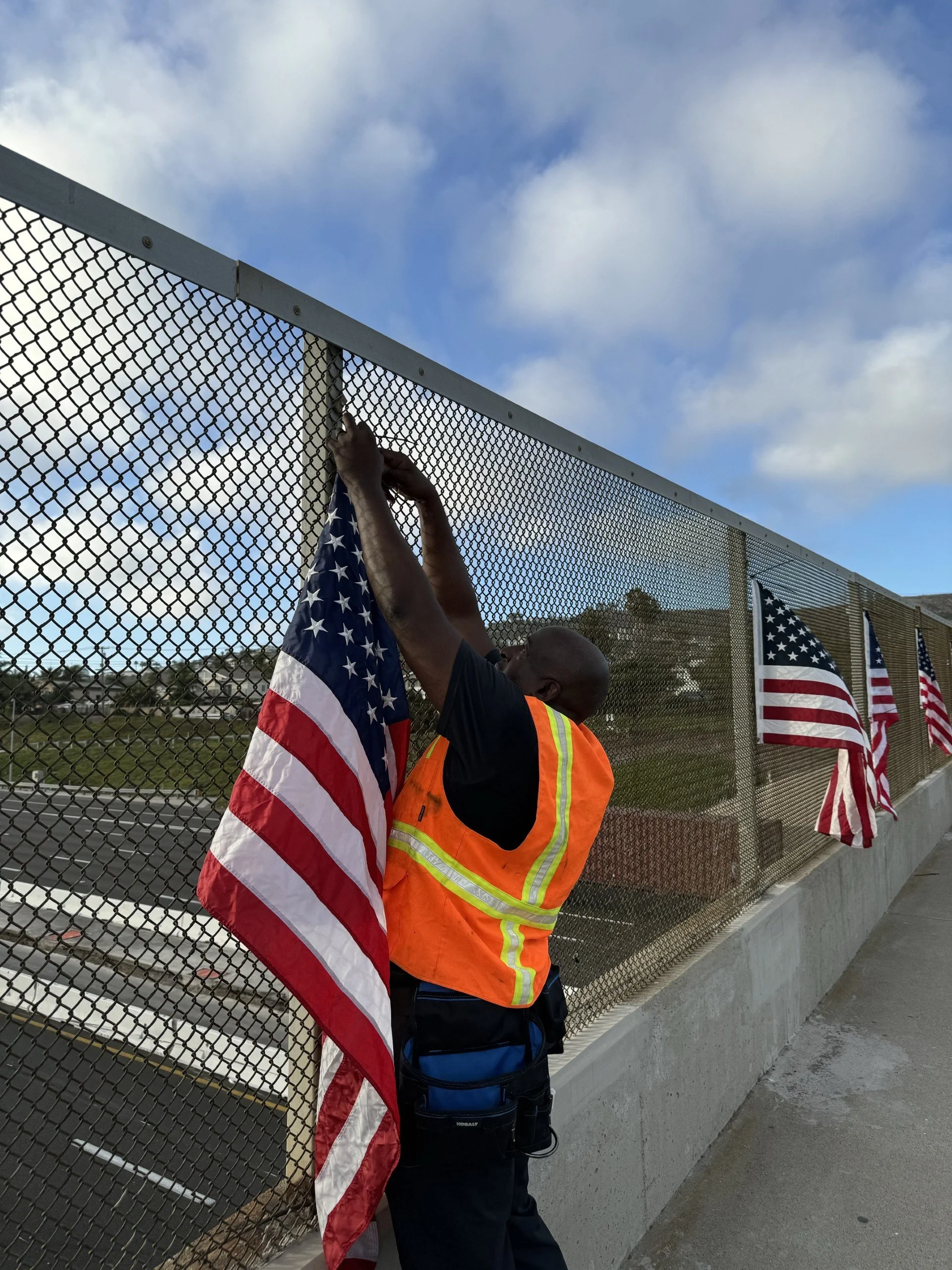 A man in an orange safety vest attaches an American flag to a chain-link fence, with multiple other American flags already on the fence, under a partly cloudy sky.