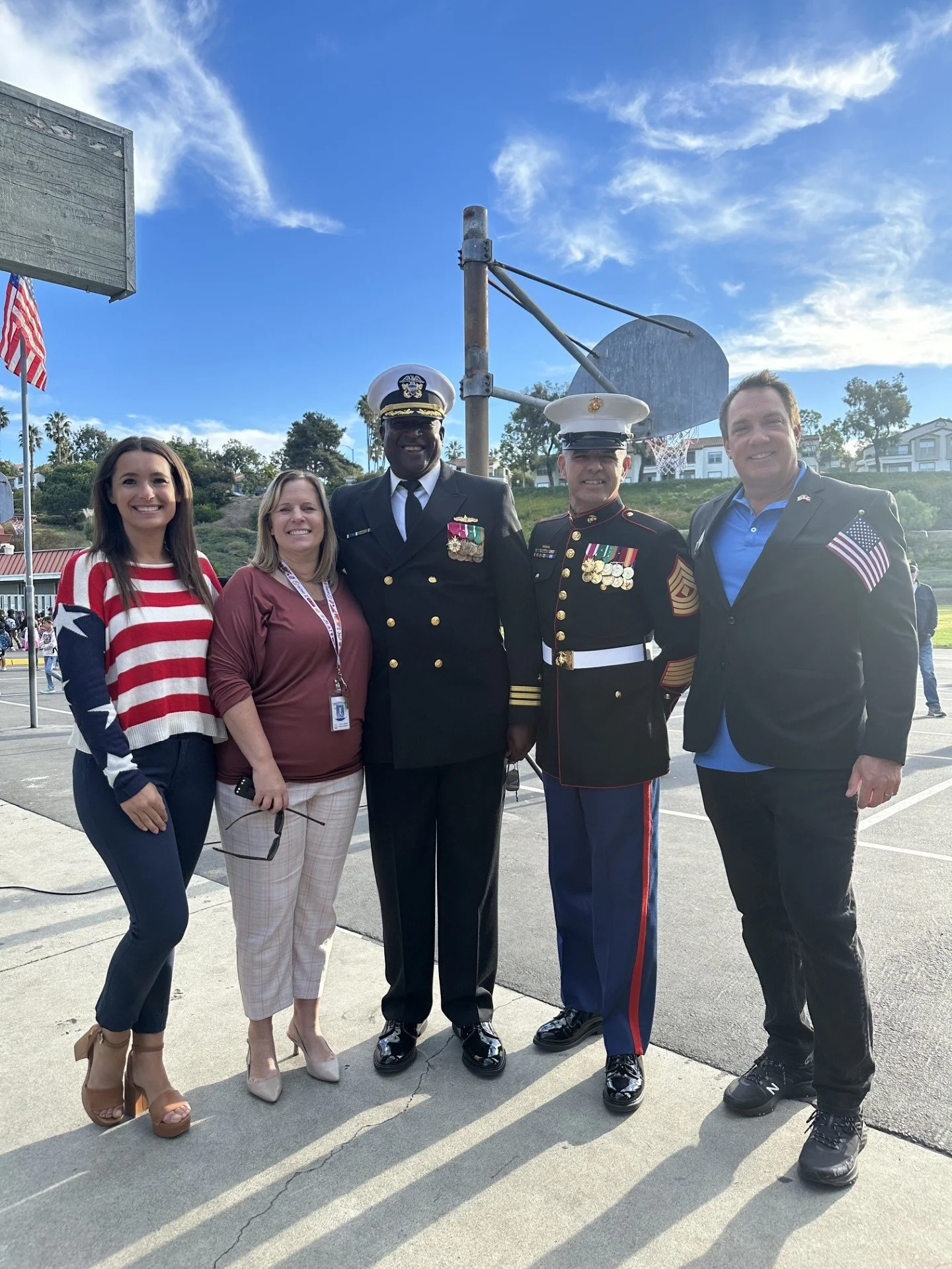 Group of five people standing together outdoors, including two men in military dress uniforms with medals, and three women, one in patriotic clothing. They are smiling in front of a basketball hoop and a partly cloudy sky.