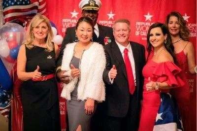 Group of five people at a celebration with balloon decorations, holding glasses and giving thumbs up, after a formal event or award ceremony.