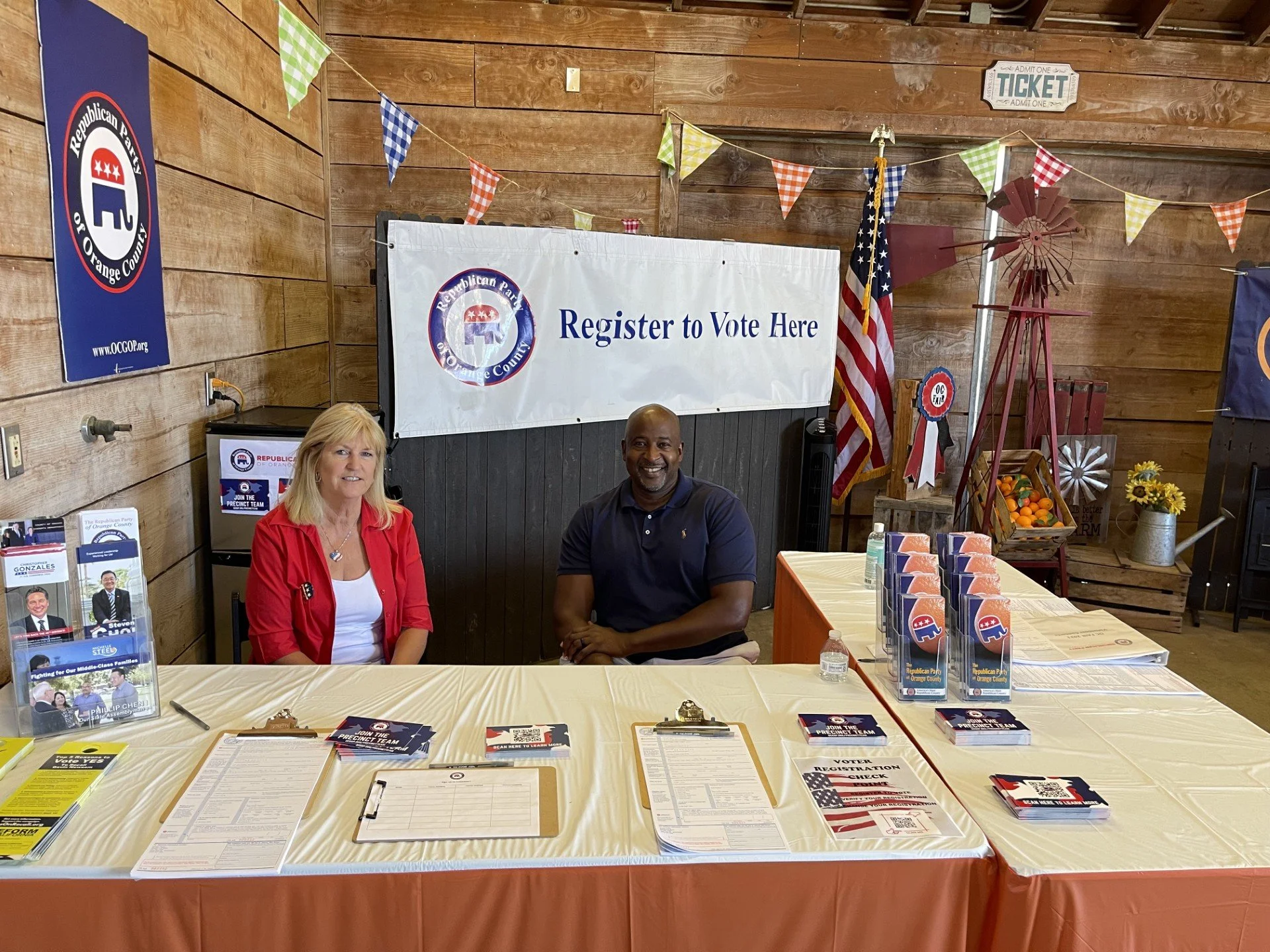 A man and a woman seated behind a table at a voting registration booth. The table has political pamphlets, a clipboard, and bottled water. The background includes a large banner that says 'Register to Vote Here', an American flag, and various politic