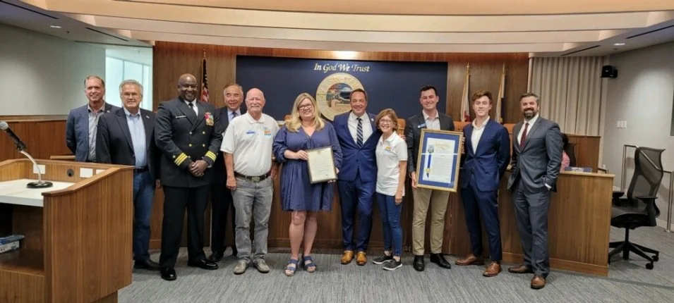 Group of people standing in a council chamber, some holding certificates, with a blue banner in the background that says 'In God We Trust' and features a seal of a government or military organization. Flags are also present.