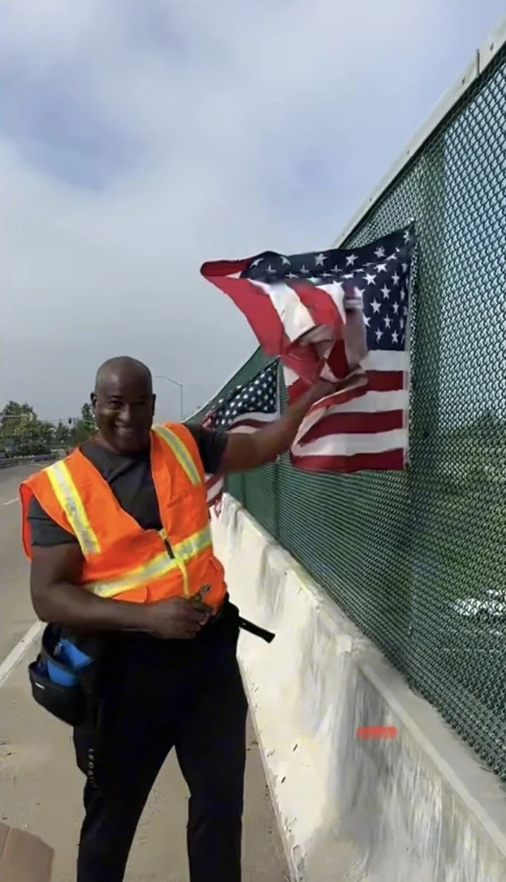 A man in a safety vest is standing by a fence, holding an American flag that is draped over the fence, with a background of a cloudy sky.