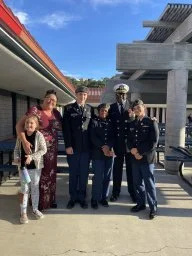 A group of five people, including two adults and three children, standing outdoors near a modern architectural building and a train or tram. The adults are dressed casually, and one is in a police uniform. They are smiling and posing for the photo.