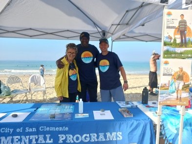 Three people standing under a canopy at a community event on the beach, with pamphlets and a tablecloth for City of San Clemente mental programs, and a promotional poster to the side.