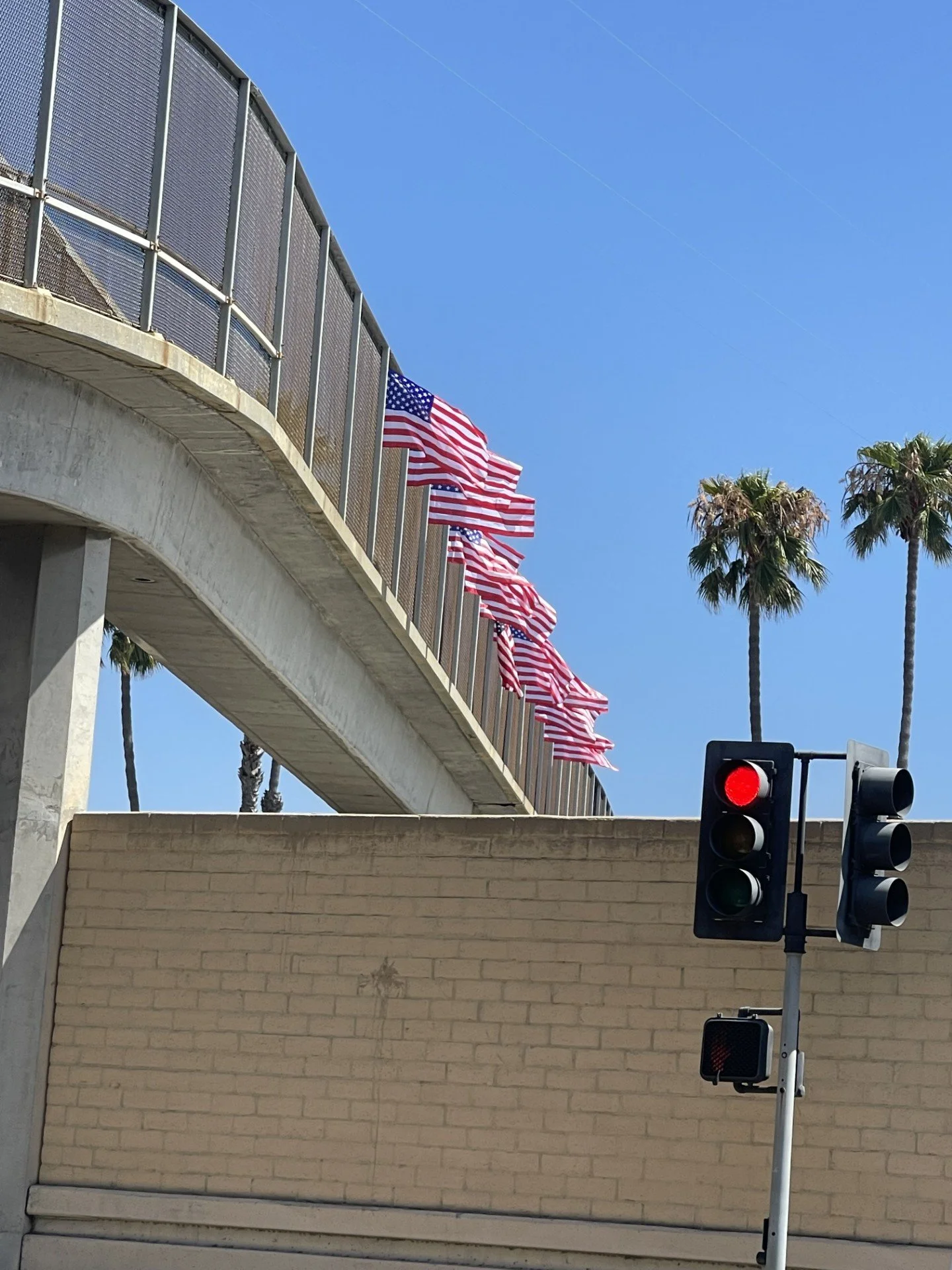 Multiple American flags hanging from a curved bridge structure alongside a traffic light with a red signal, palm trees in the background, and a clear blue sky.