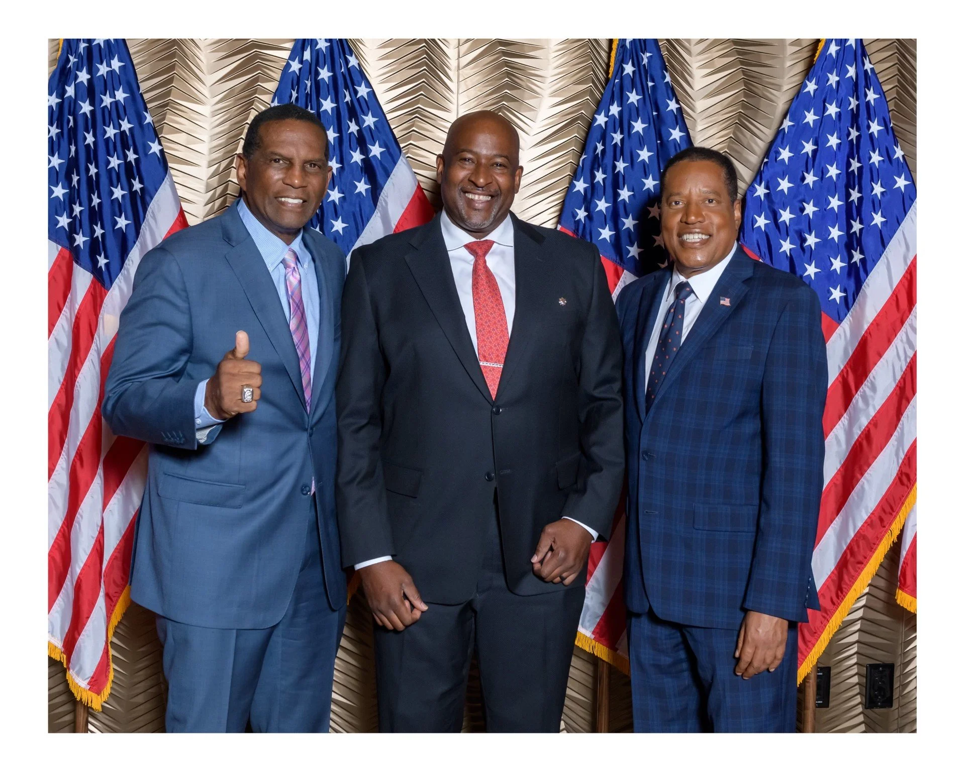 Three men in suits standing in front of U.S. flags, smiling for the photograph. The man on the left is giving a thumbs up.
