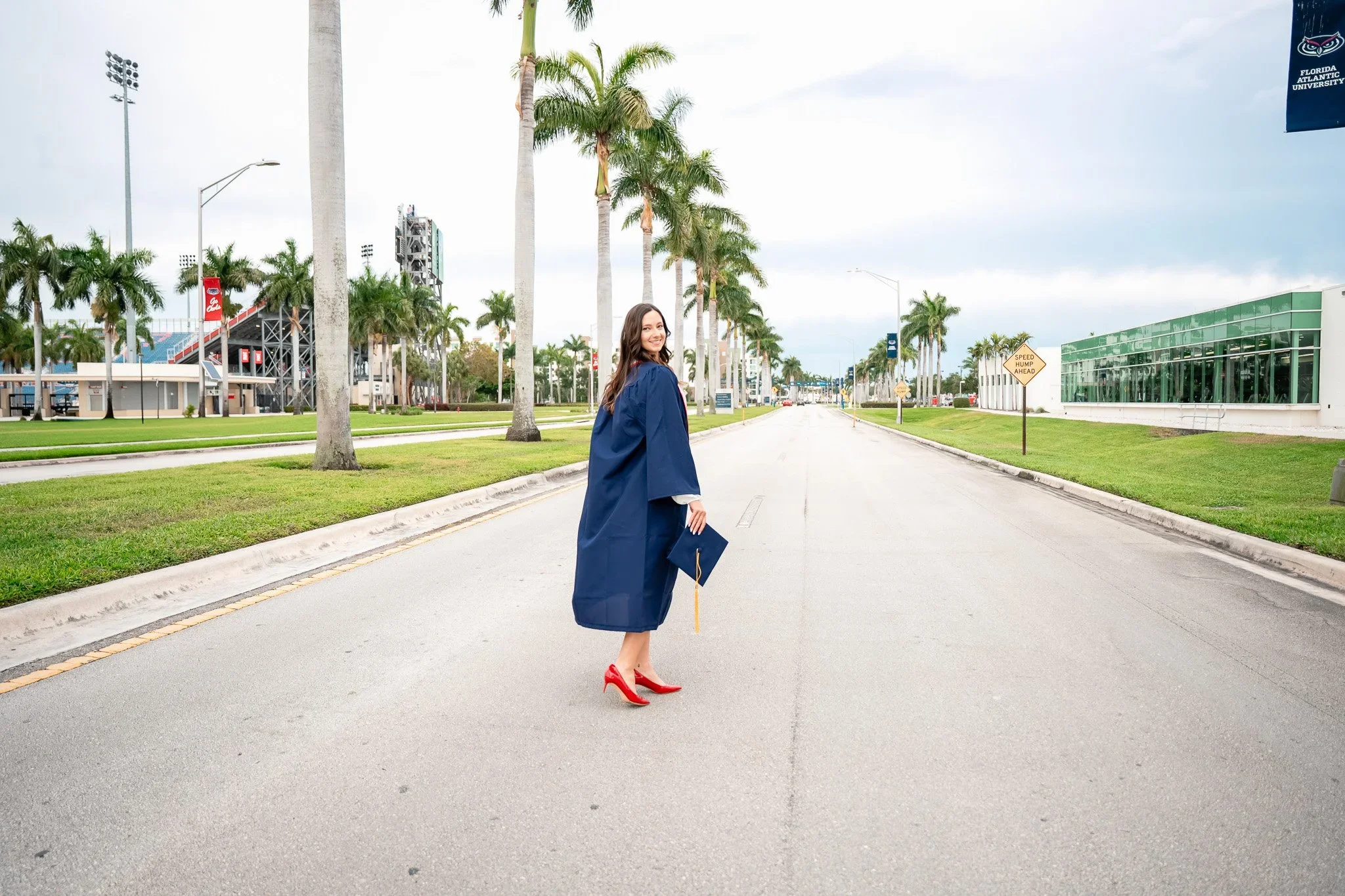 FAU graduate in a navy gown and red heels holding her cap on a palm-lined campus road