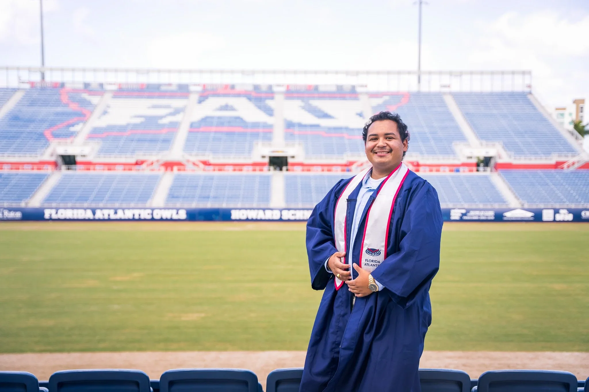 FAU graduate posing on the football field with the stadium seating behind him