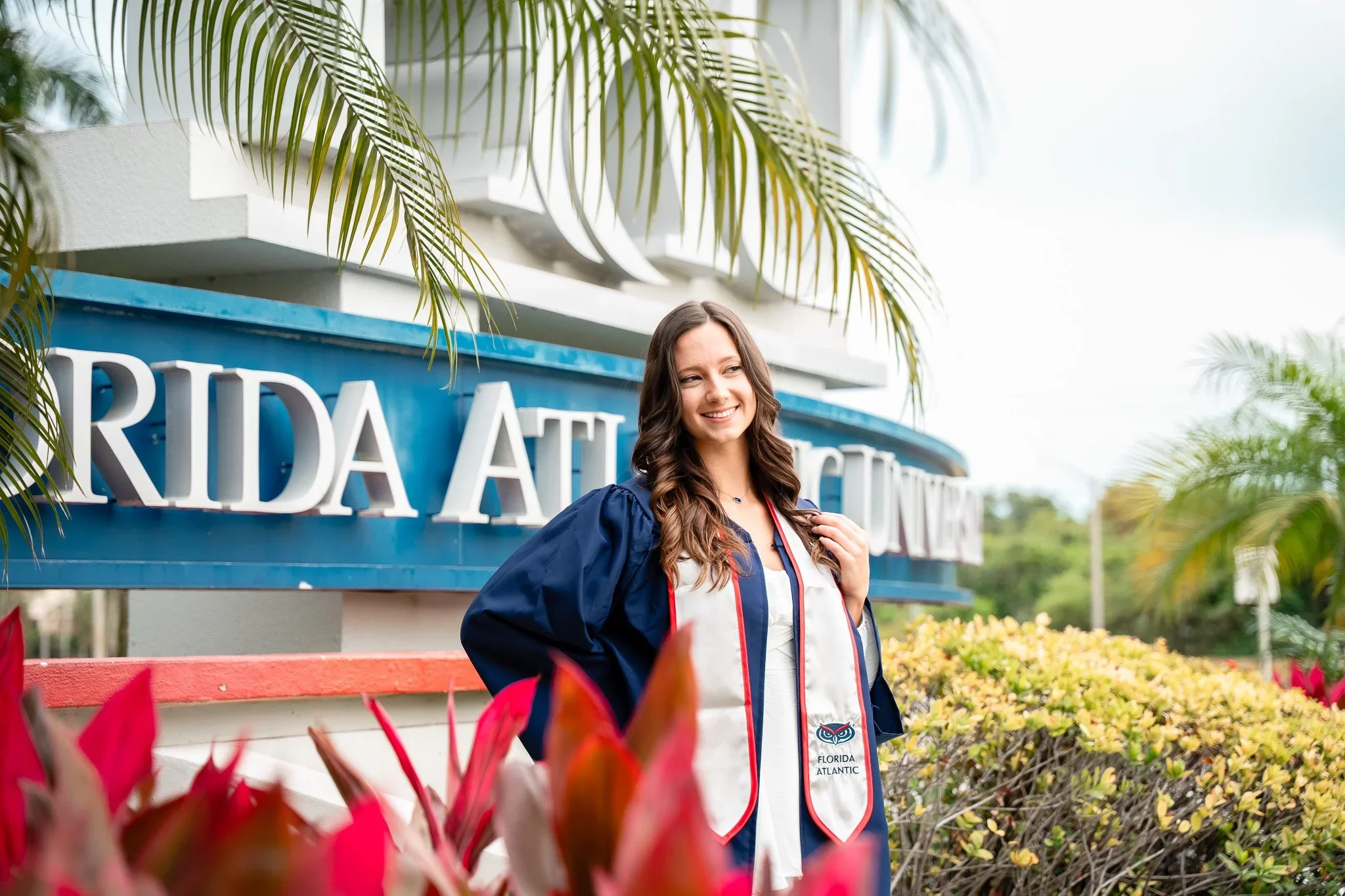 FAU graduate in a navy gown and stole posing by the Florida Atlantic University sign