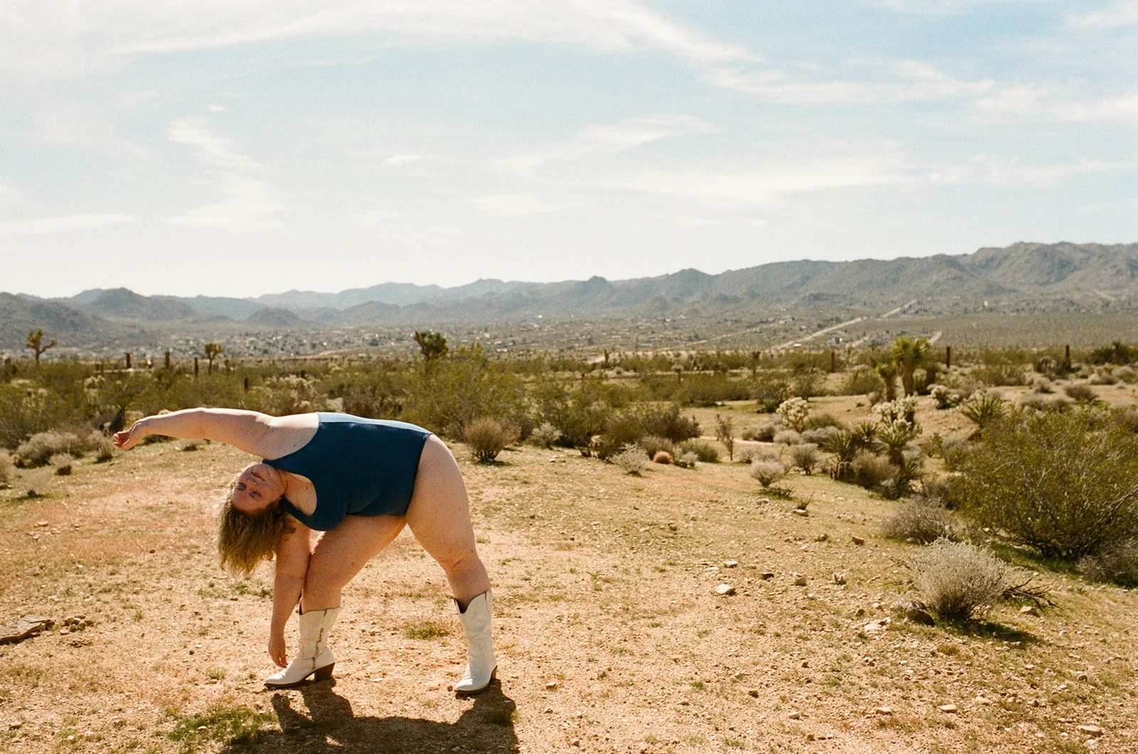 woman in blue bodysuit and white cowboy boots bends over sideways in the desert modeling for a photography workshop