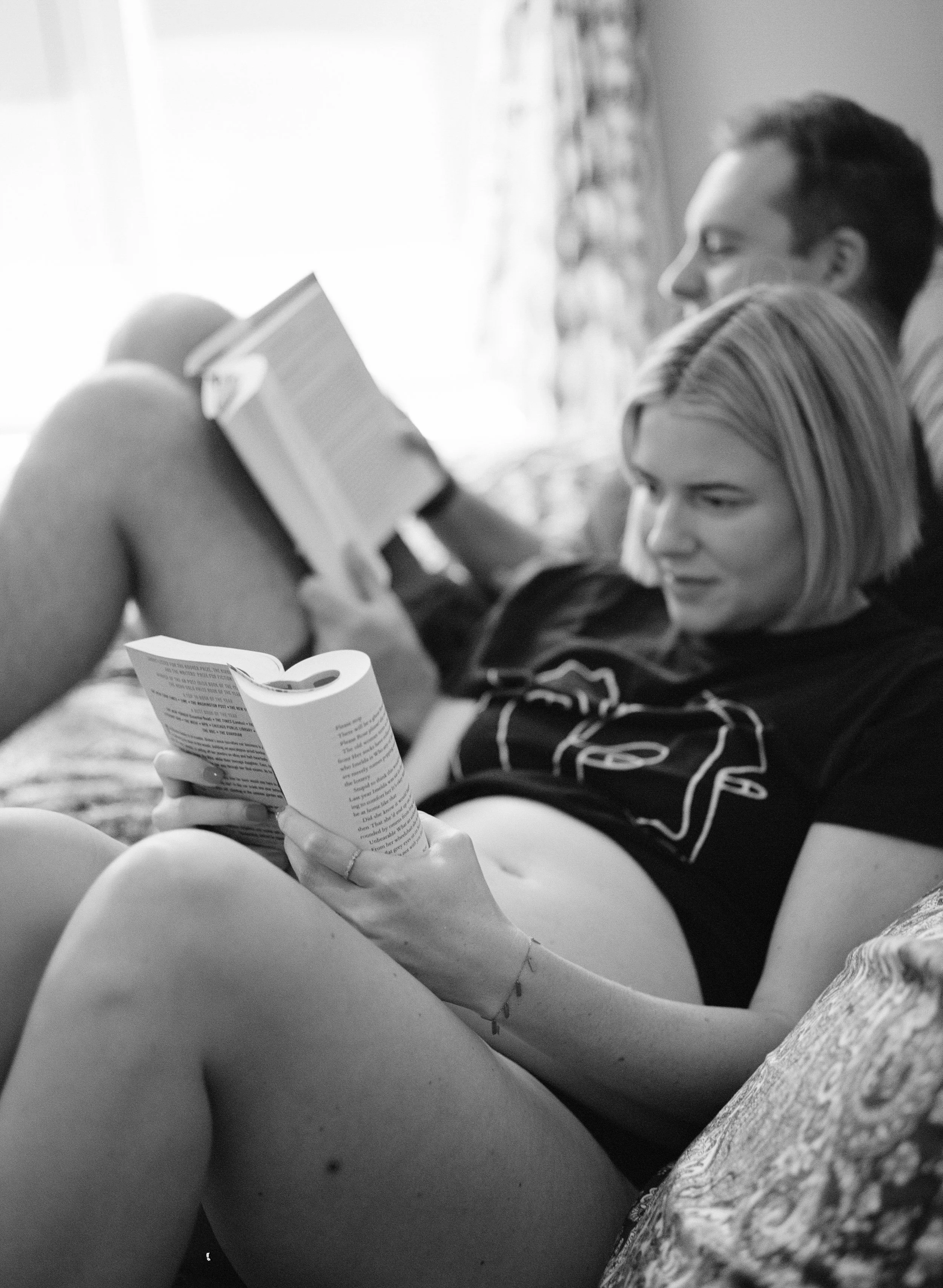 Pregnant woman and her spouse lounge while reading books during their family photo session. 