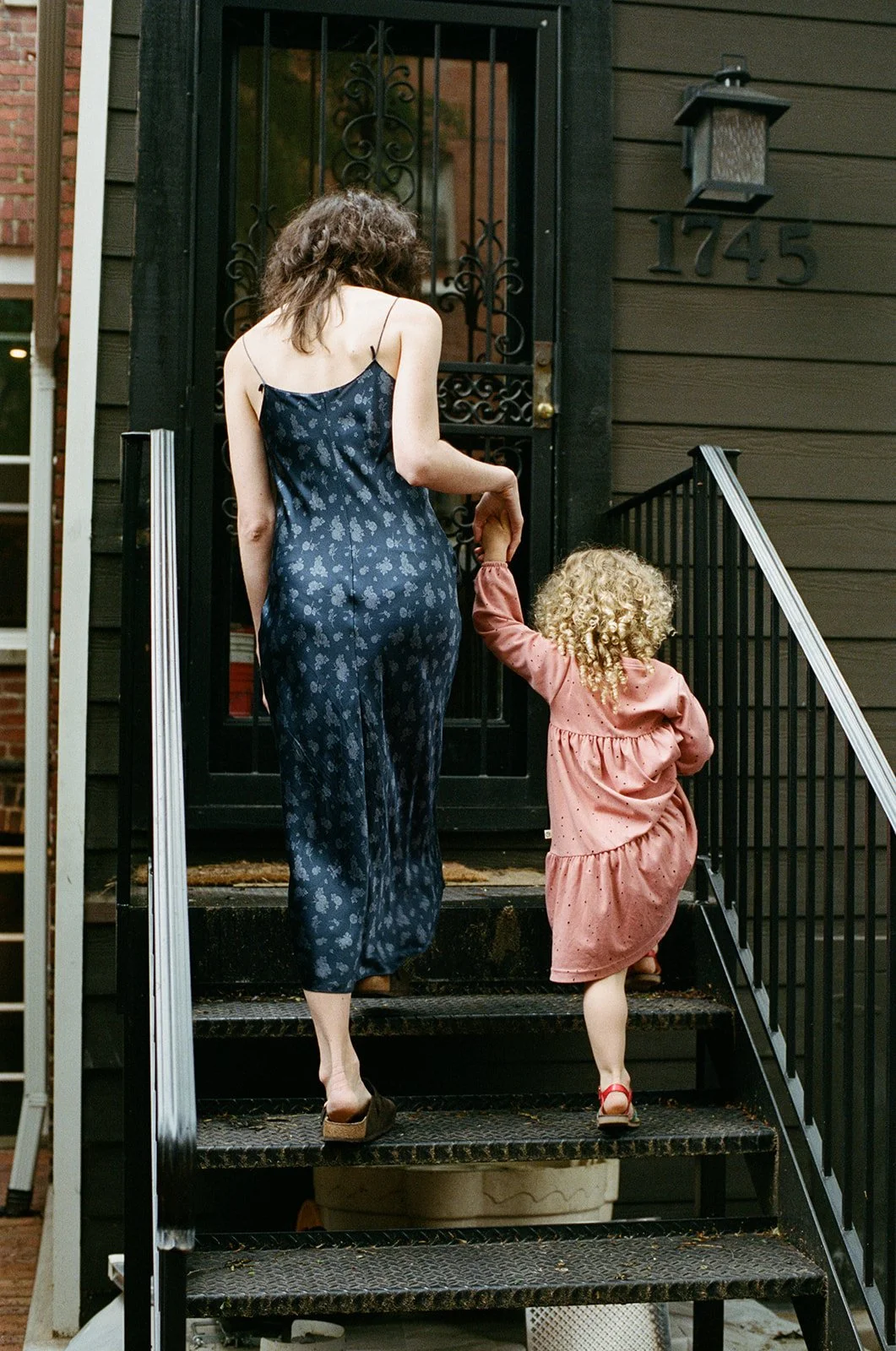 a mother and child are walking up the steps to their front door in unison, both with curly hair wearing dresses 