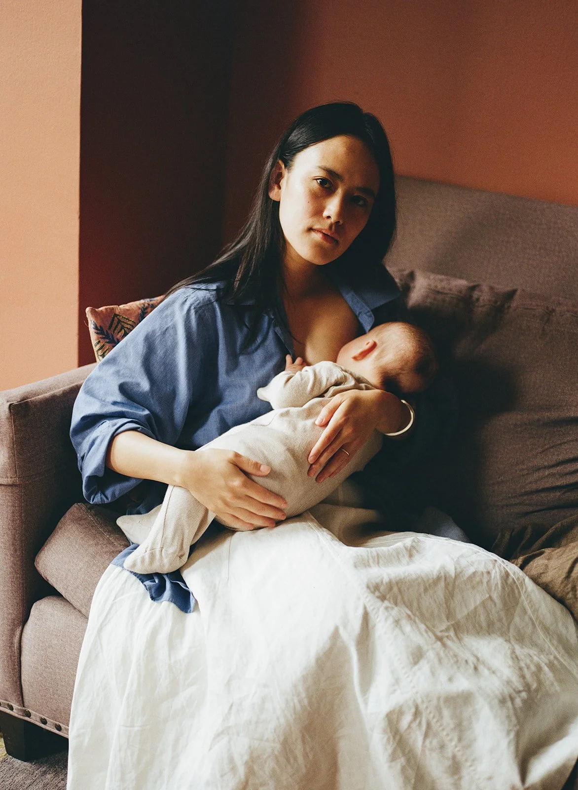 mother sits in a chair with directional light from the window to her right while posing for a photo breastfeeding her infant child 