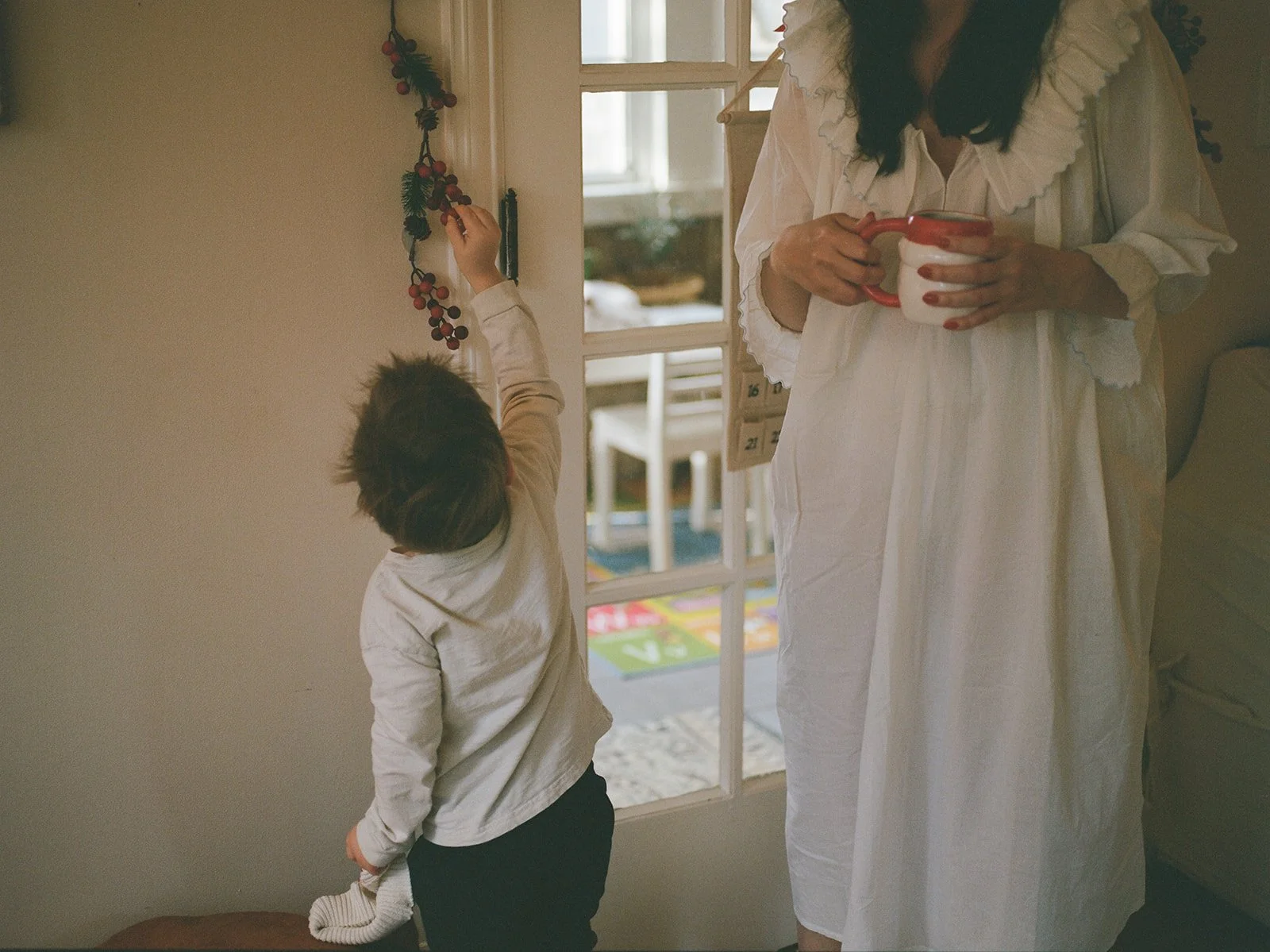 mother wearing a white ruffled dress holds her coffee cup while standing next to a boy who is admiring holiday decorations