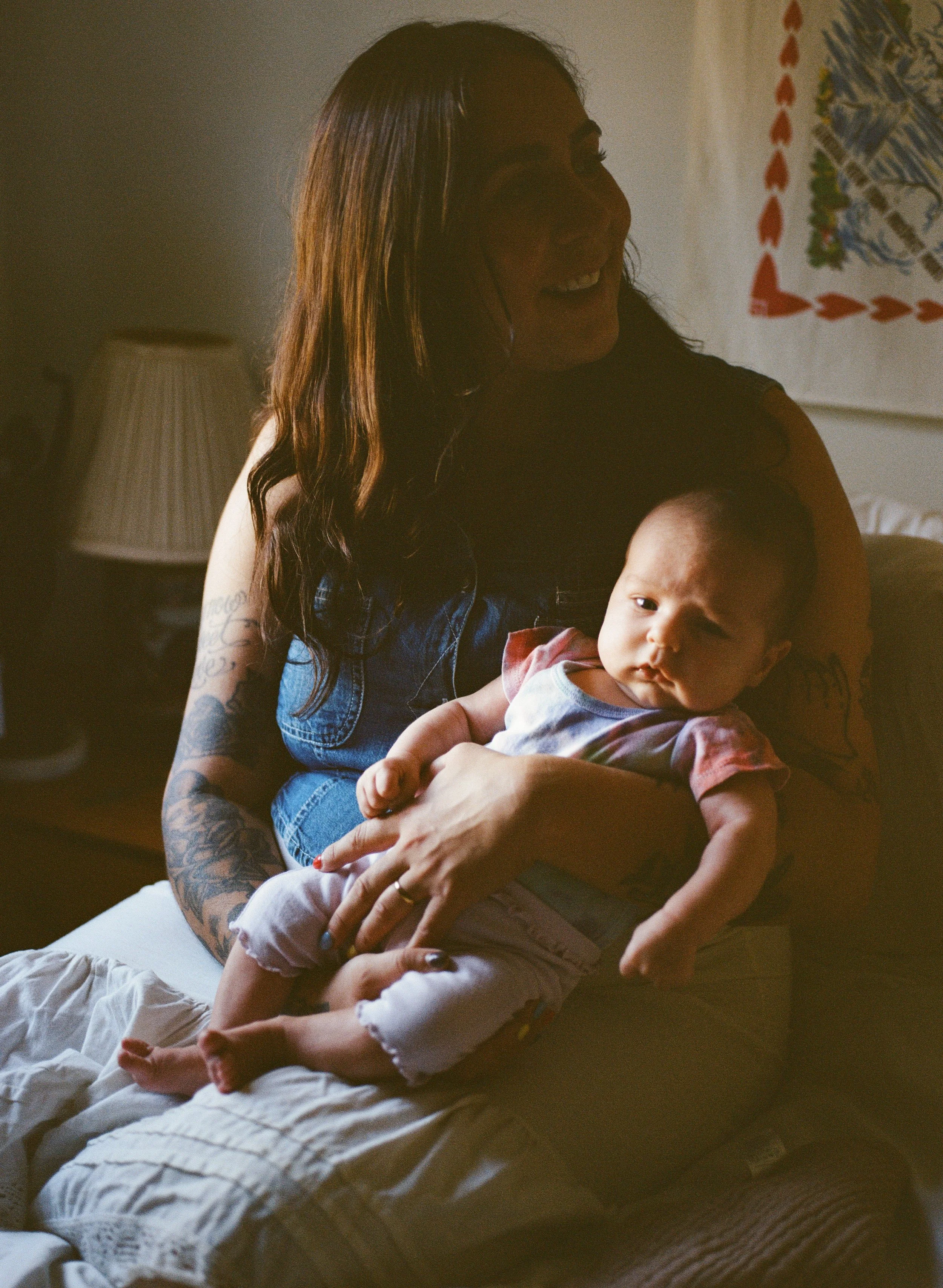 A mother sits in shadow holding an infant highlighted by chiaroscuro style lighting from a window to the mother's right. 