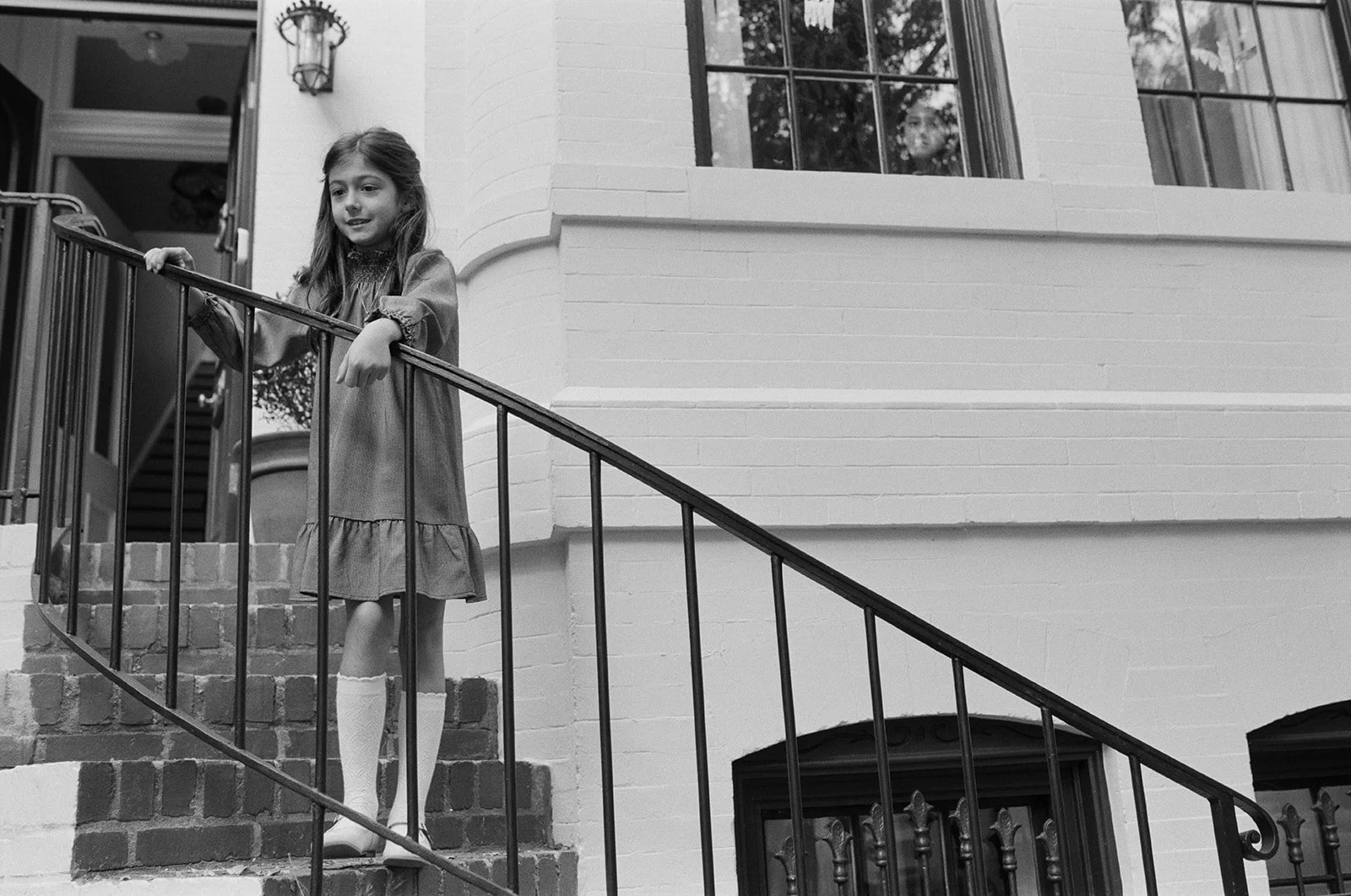 a black and white on film image in which a young girl stands at the railing, halfway down from the steps to her front door 