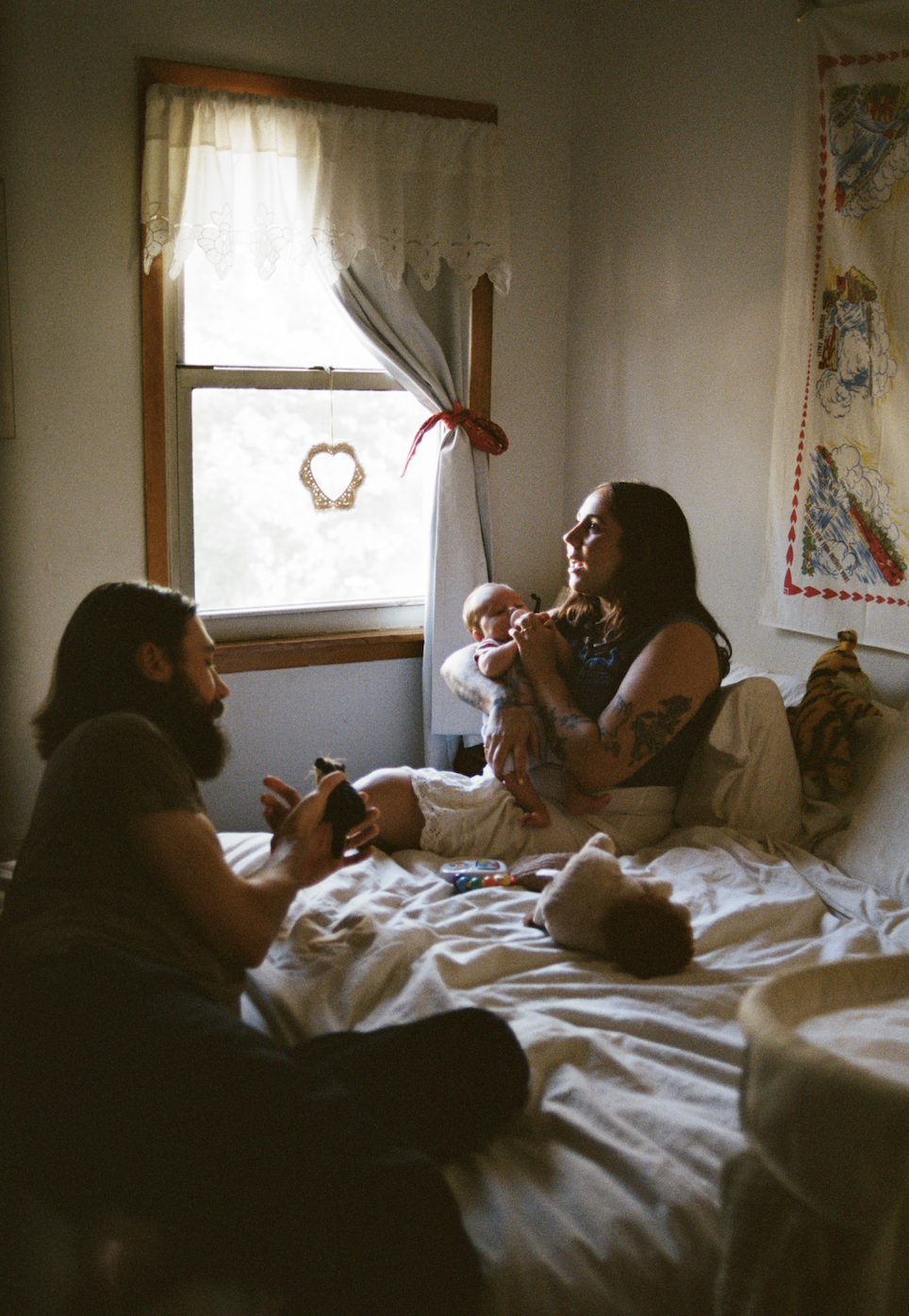 A mother and infant sit together on the bed illuminated by window light while the father sits further back in the shadows of the foreground. 