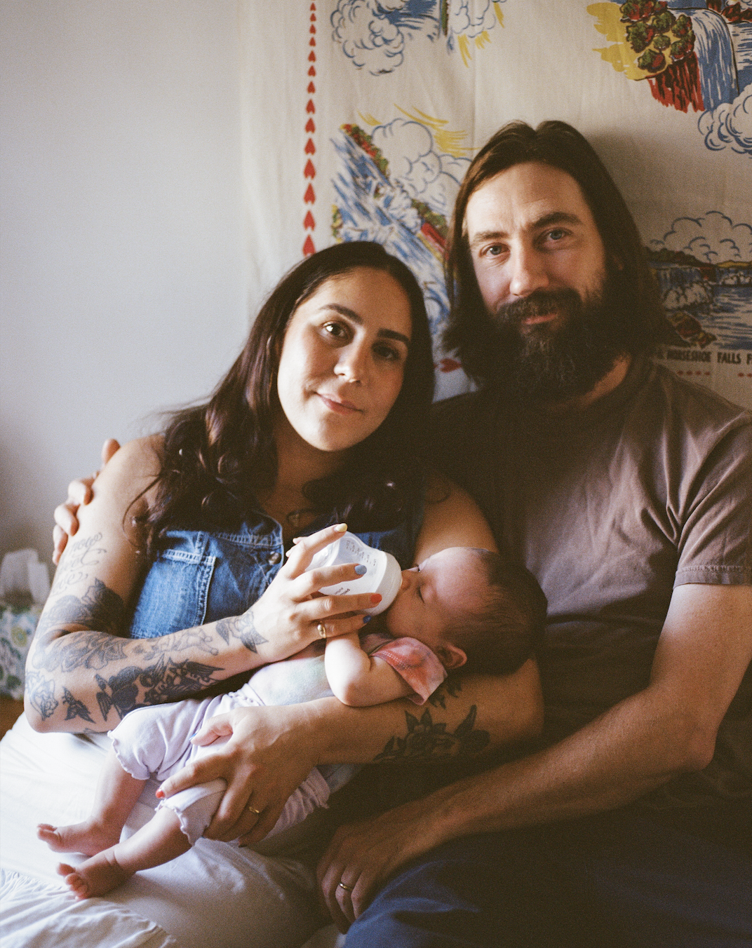 A family portrait with the parents smiling at the camera while the feed their newborn a bottle during an in home newborn photography session with film photographer Rachel Larsen Weaver. 