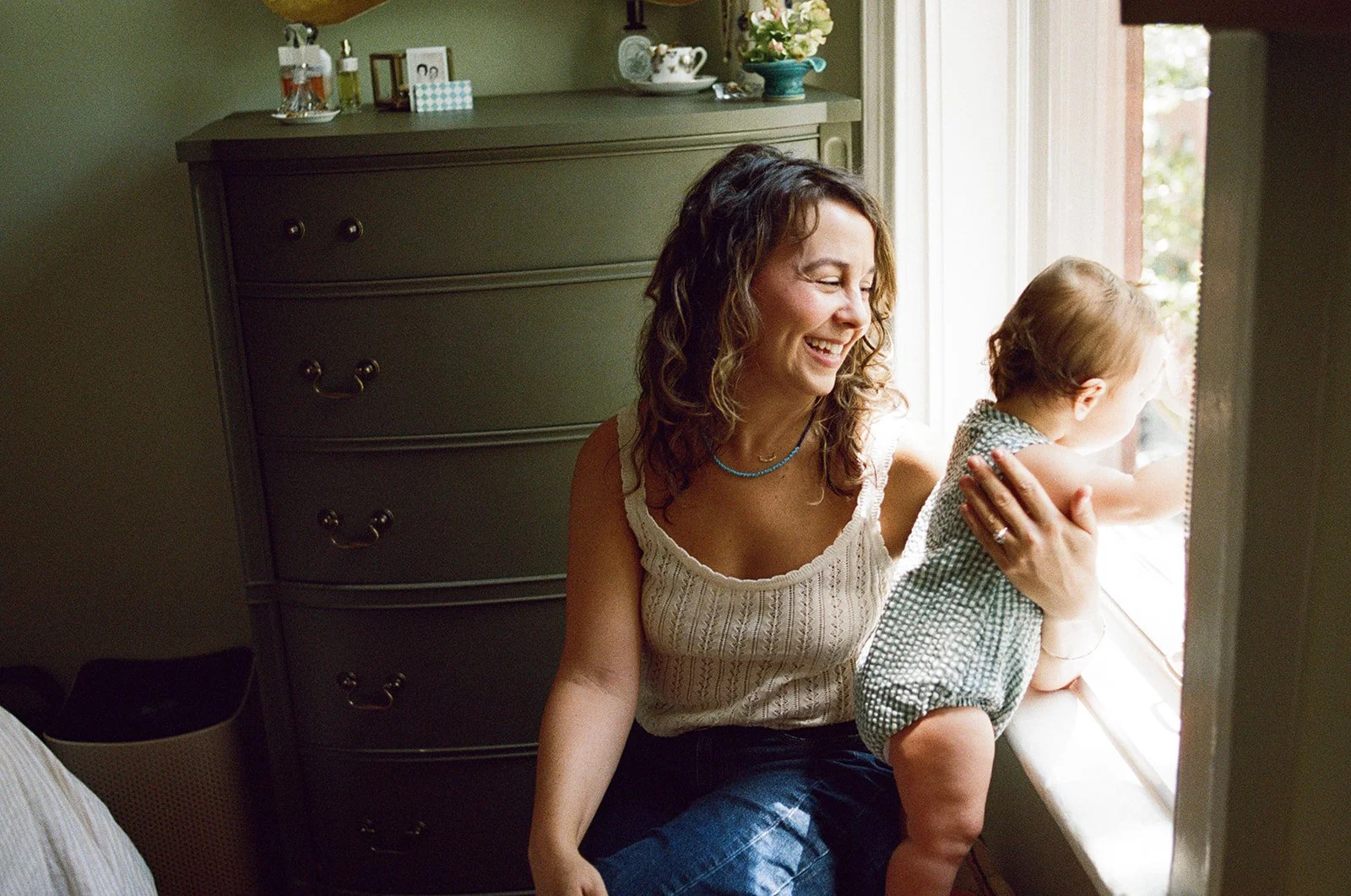 a mother laughs at her baby looking out the window during a family photo session in their home.