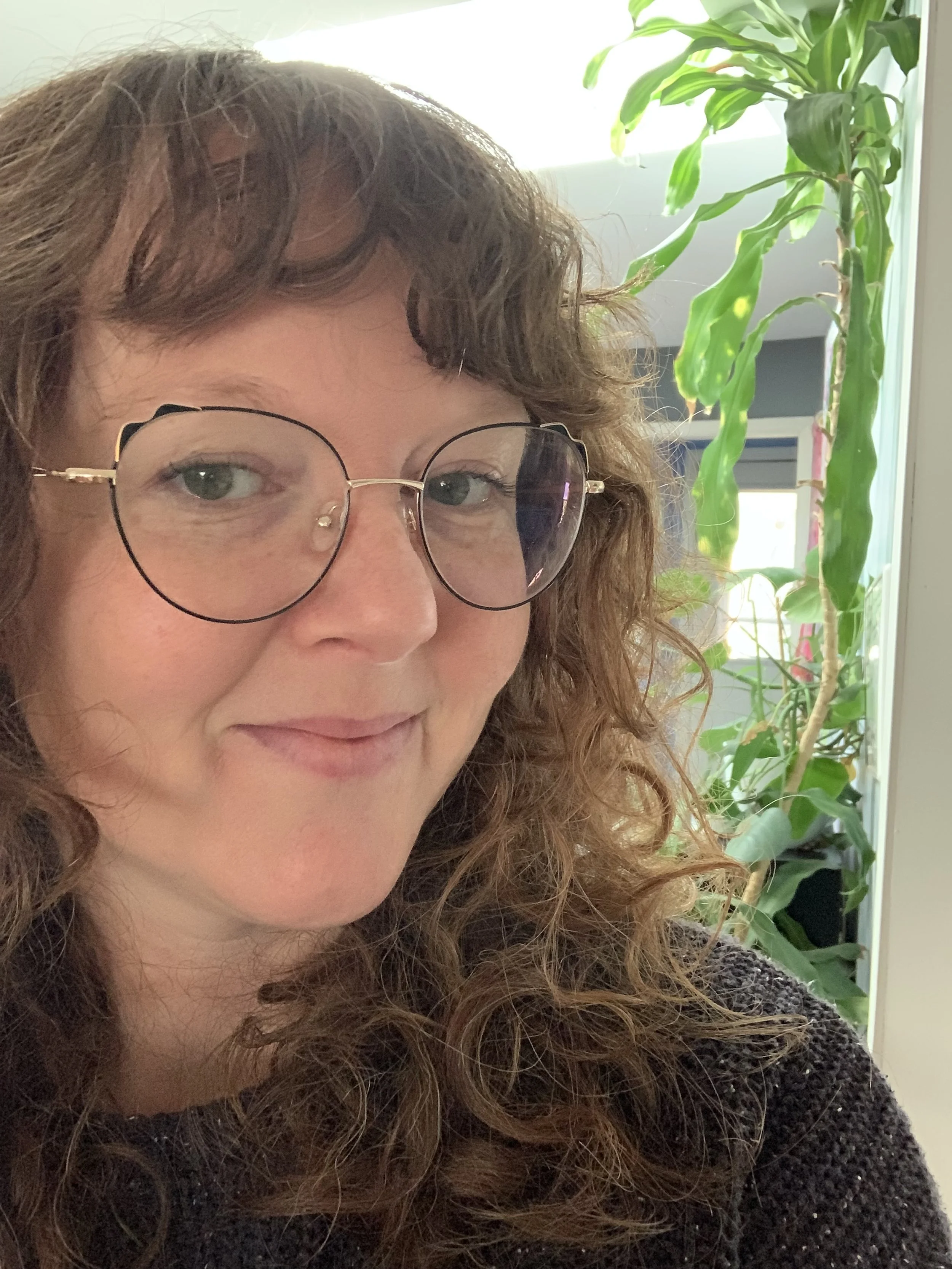 Woman with curly hair and large glasses smiling, indoors with plants in the background.