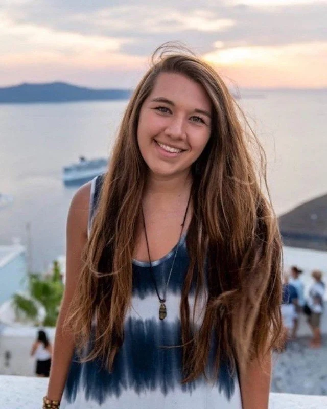 A woman with long hair smiling in front of a scenic ocean view at sunset.