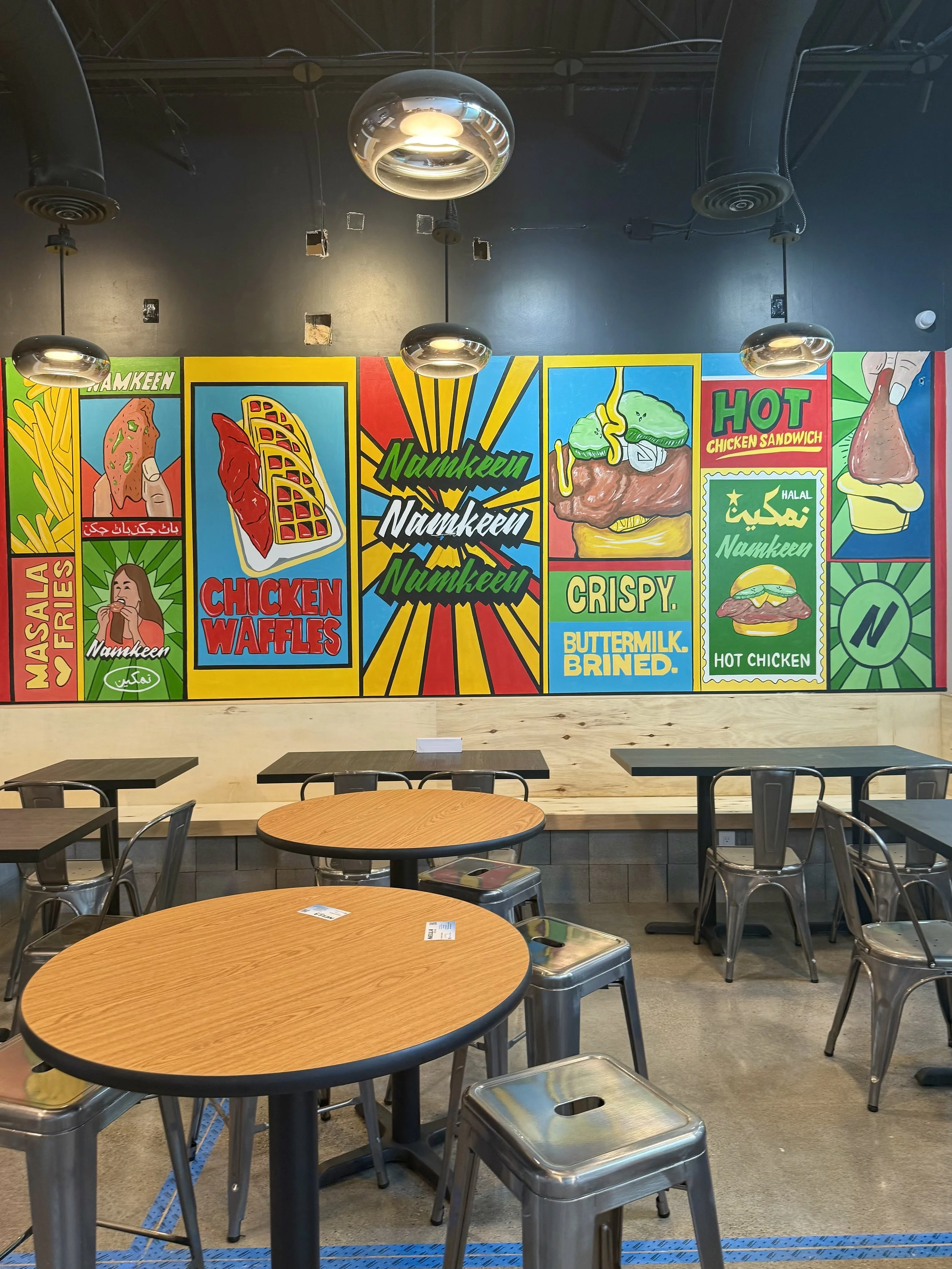Interior of a fast food restaurant with wooden tables, metal chairs, and a colorful mural on the wall advertising chicken waffles, hot chicken, and other menu items with vibrant graphics.