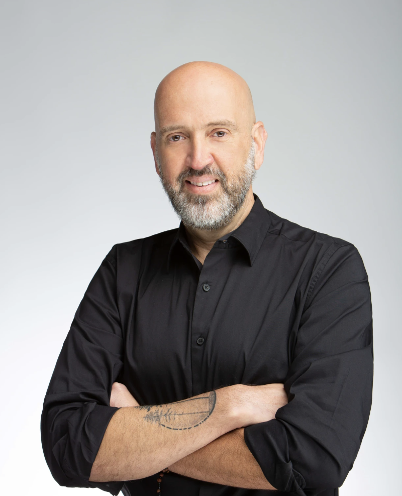 Bald man with beard in black shirt, arms crossed, tattoo on forearm, smiling.