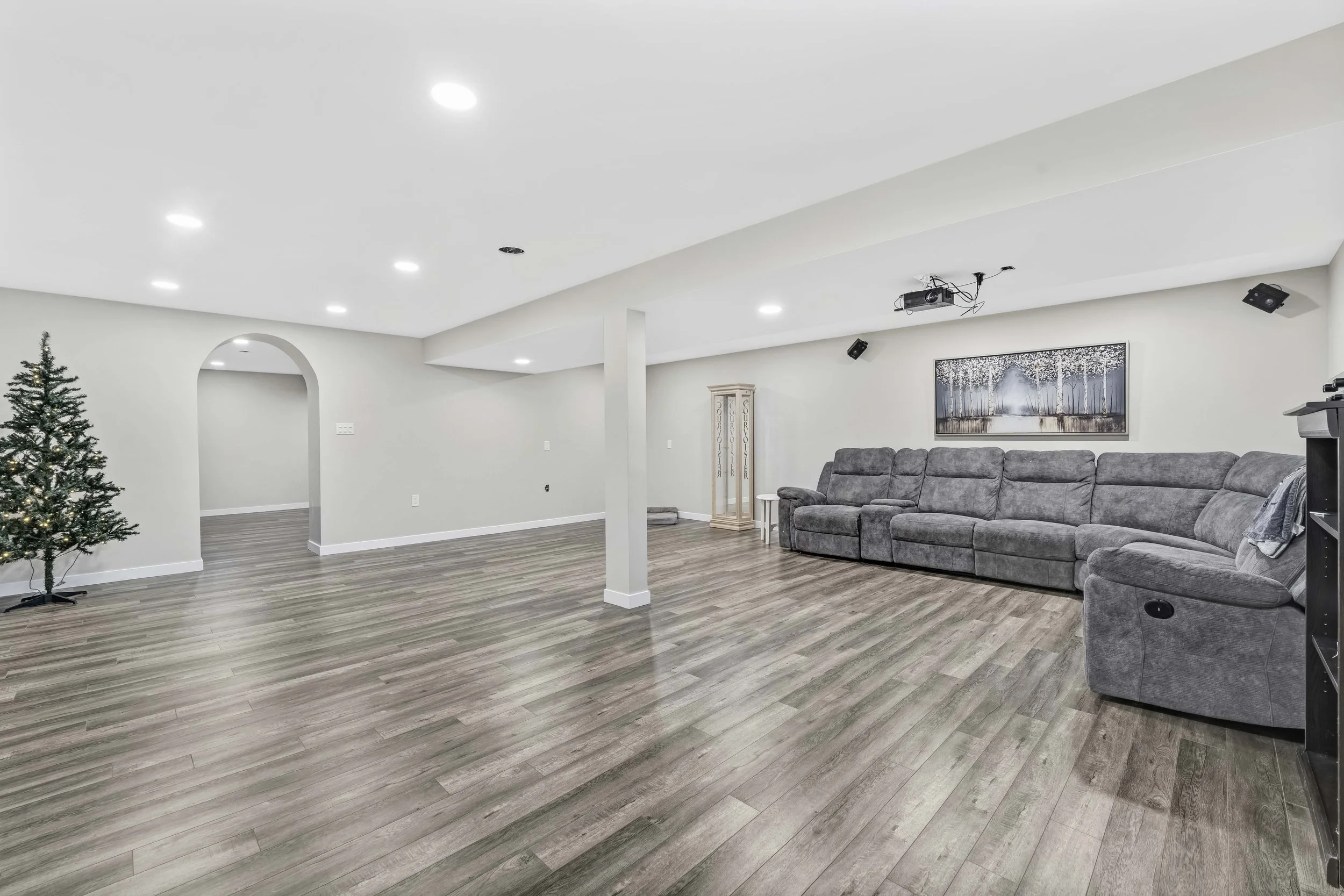 NV3D Ottawa Real Estate Photography Empty basement room with a Christmas tree, hardwood floors, a large gray sectional sofa, a wall-mounted art piece, and ceiling-mounted projector and speakers.