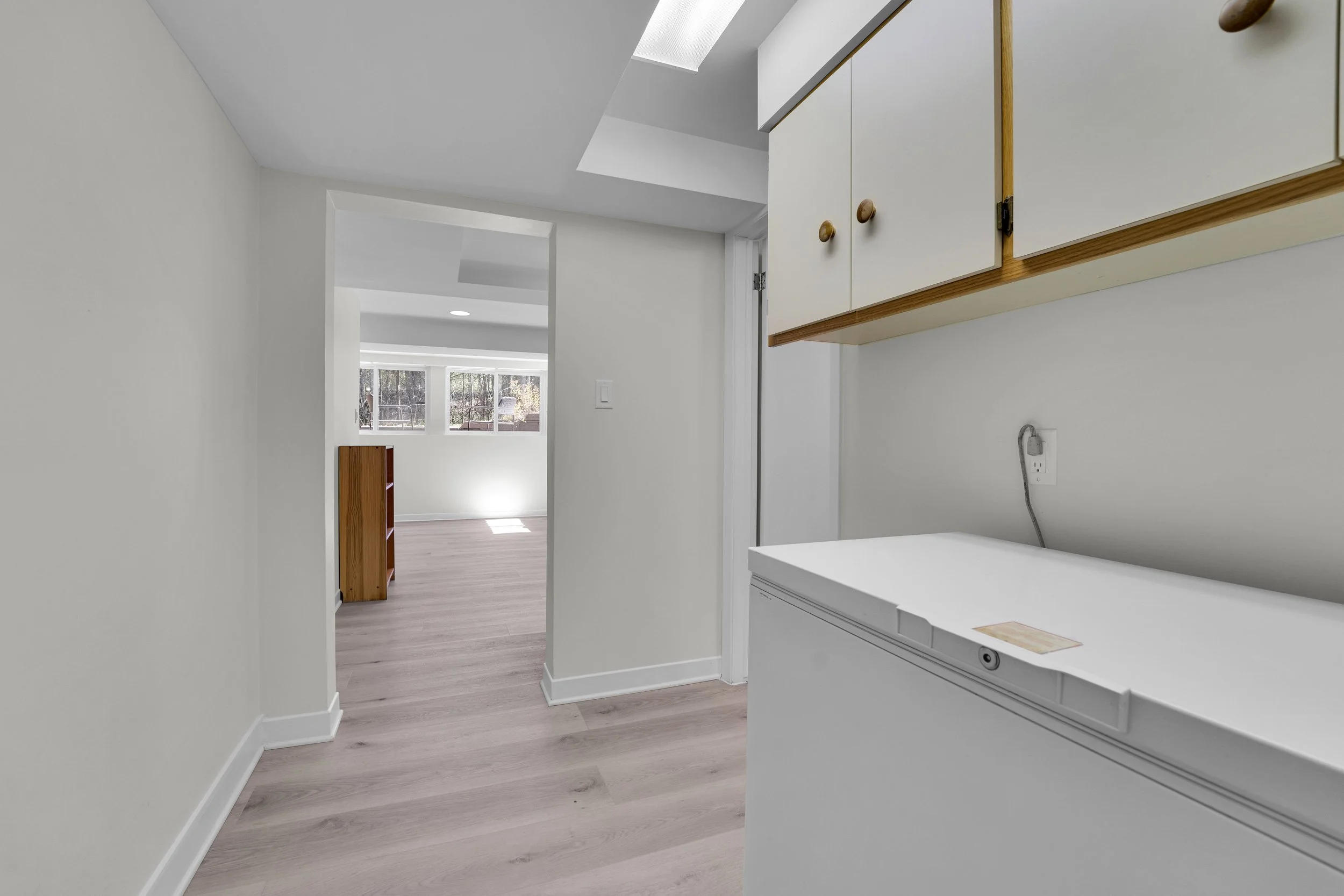 NVISION3D.ca Real Estate Media Ottawa  View of an interior space with white walls, hardwood flooring, and near a laundry area with a cabinet and a wall outlet, leading to a brightly lit room with large windows showing trees outside.