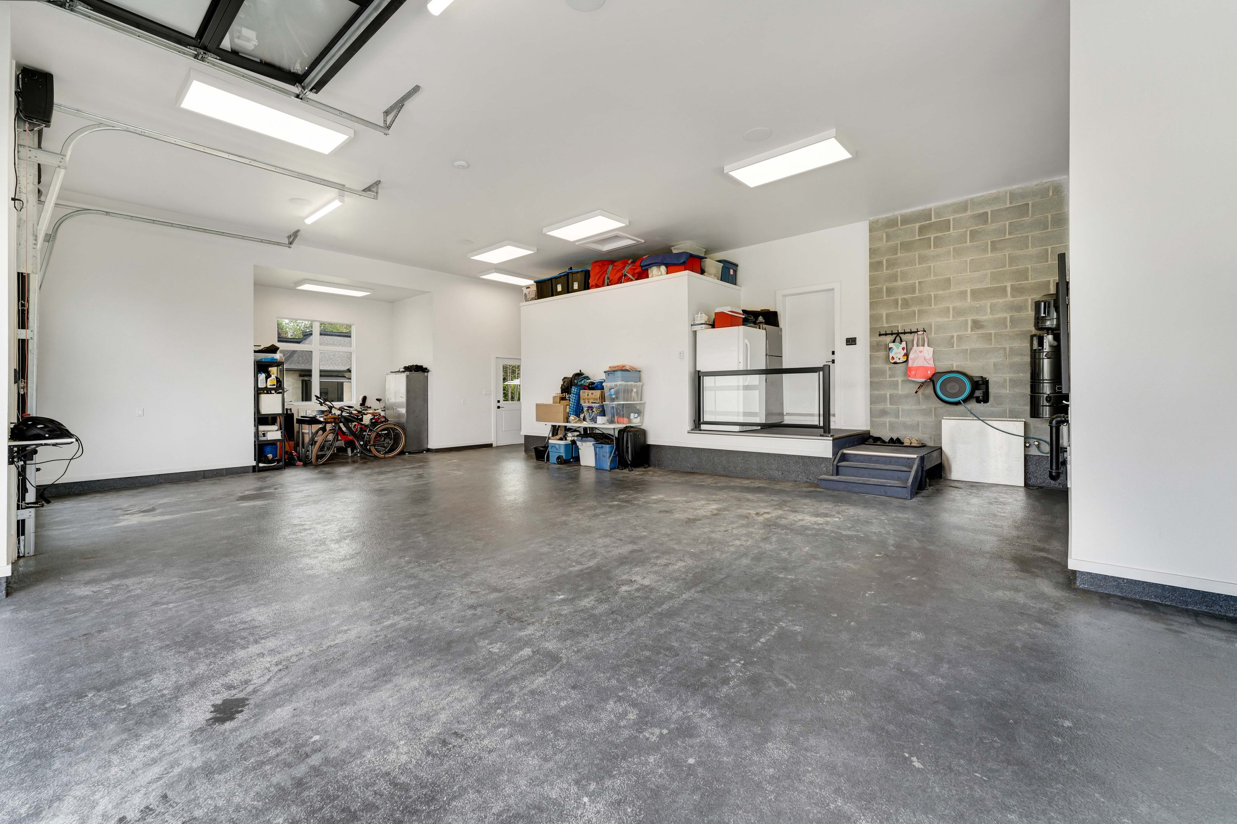 Spacious garage interior with storage shelves, bicycles, and various household items organized along the walls. The garage features overhead lighting, a concrete floor, and hooks mounted on the wall for additional storage.