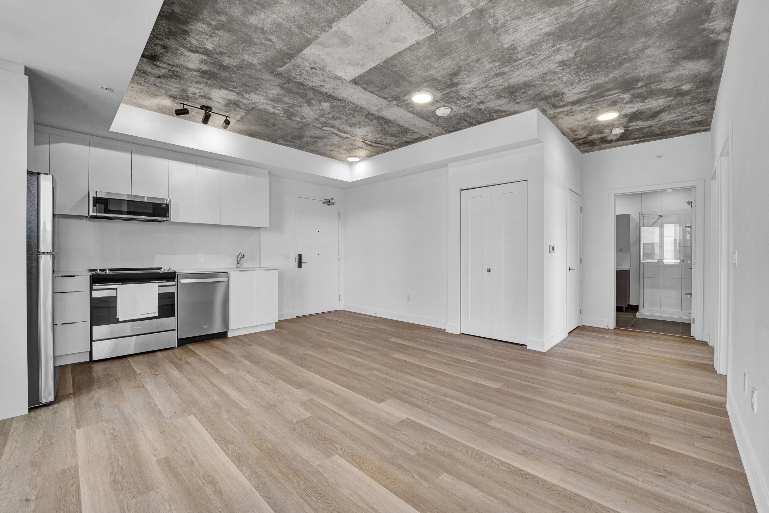 Modern kitchen with white cabinets, stainless steel appliances, and a wood floor. The ceiling has a concrete finish with recessed and track lighting.