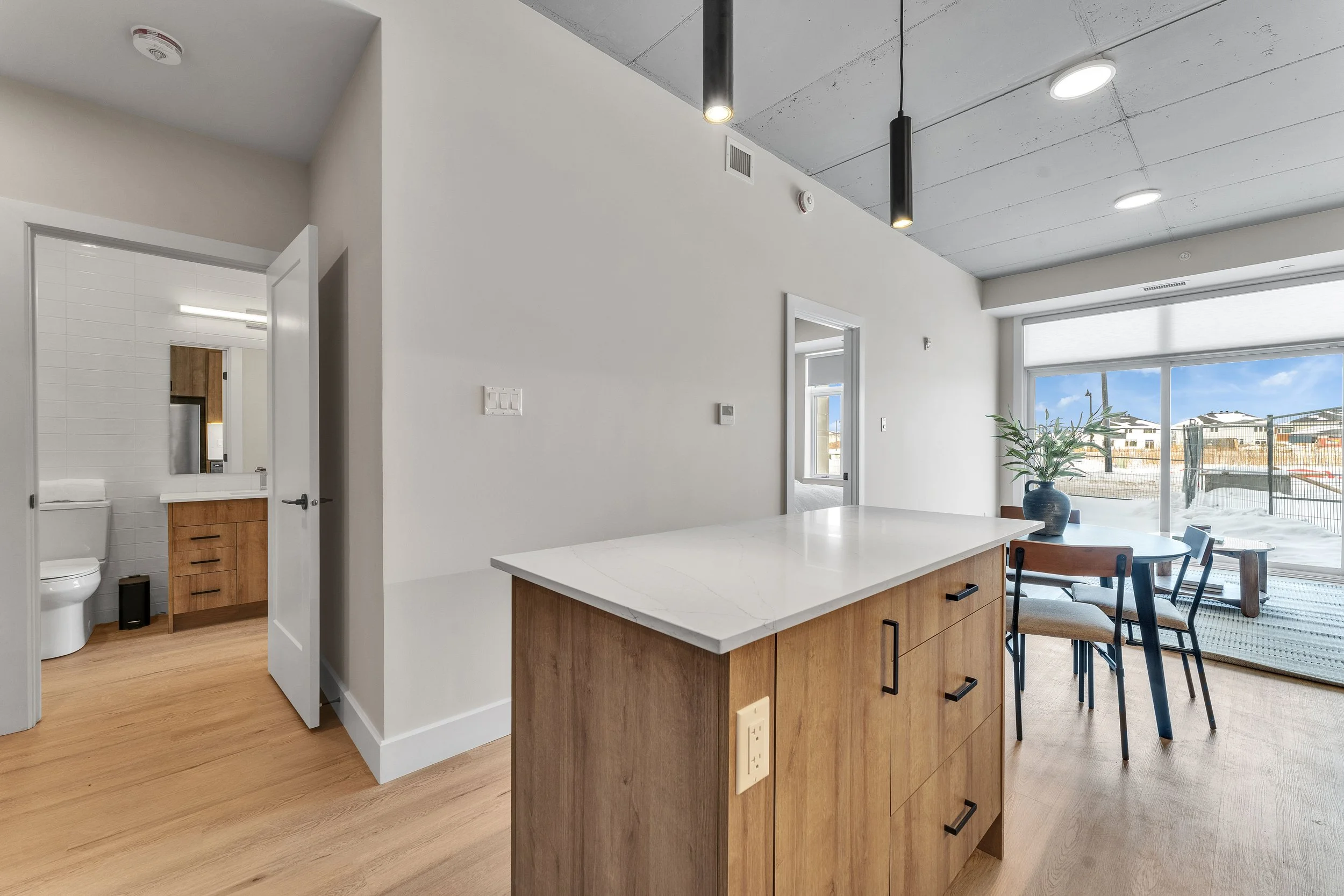 Modern open-plan apartment interior with kitchen island, dining table with chairs, large sliding glass door leading to balcony, and visible bathroom in the background.