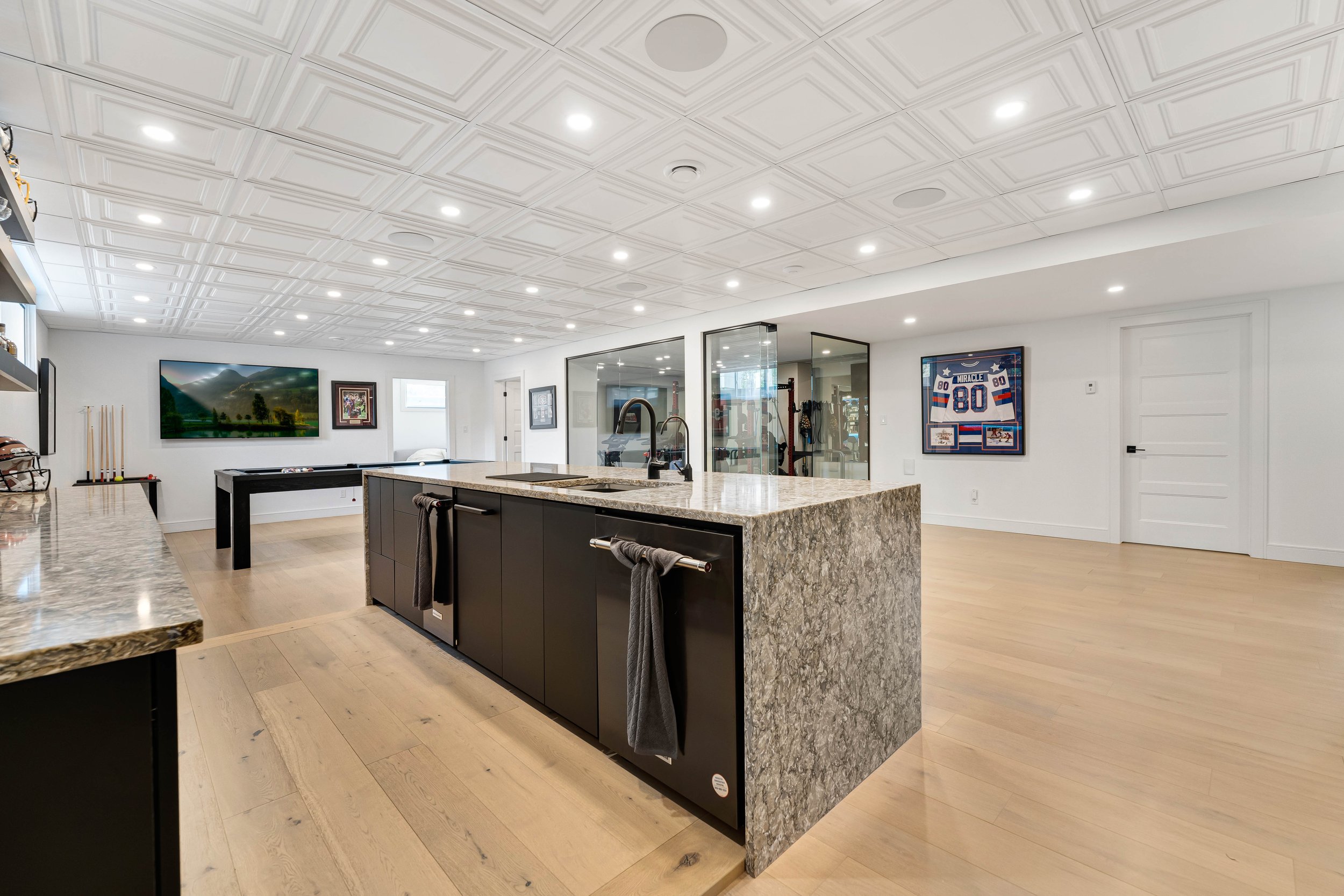 Spacious modern basement with a large kitchen island, wall decor, a pool table, and workout equipment in the background.