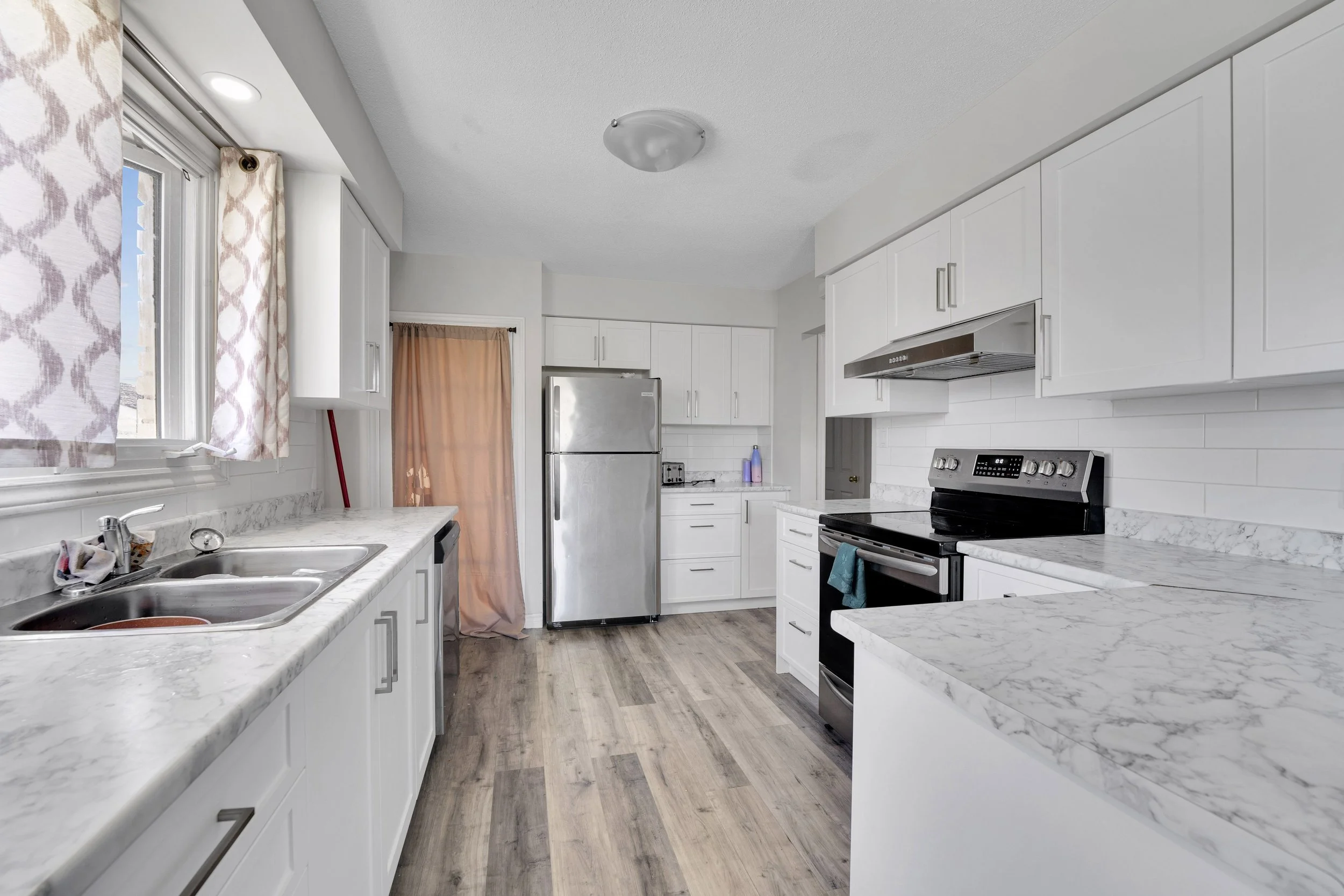 Ottawa Real Estate Media Services  A clean kitchen with white cabinets, marble countertops, a stainless steel refrigerator, black stove, and hardwood floors. There's a window with patterned curtains and a door with a pink curtain.